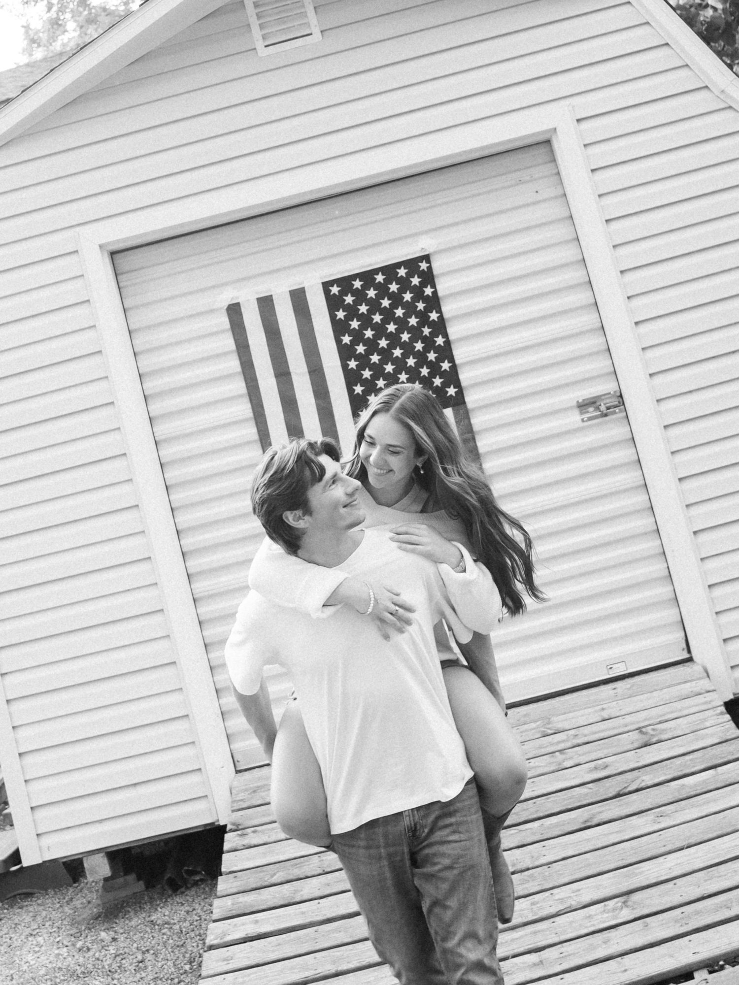 Black and white photo of a couple laughing, man giving woman a piggyback ride in front of a building with an American flag.