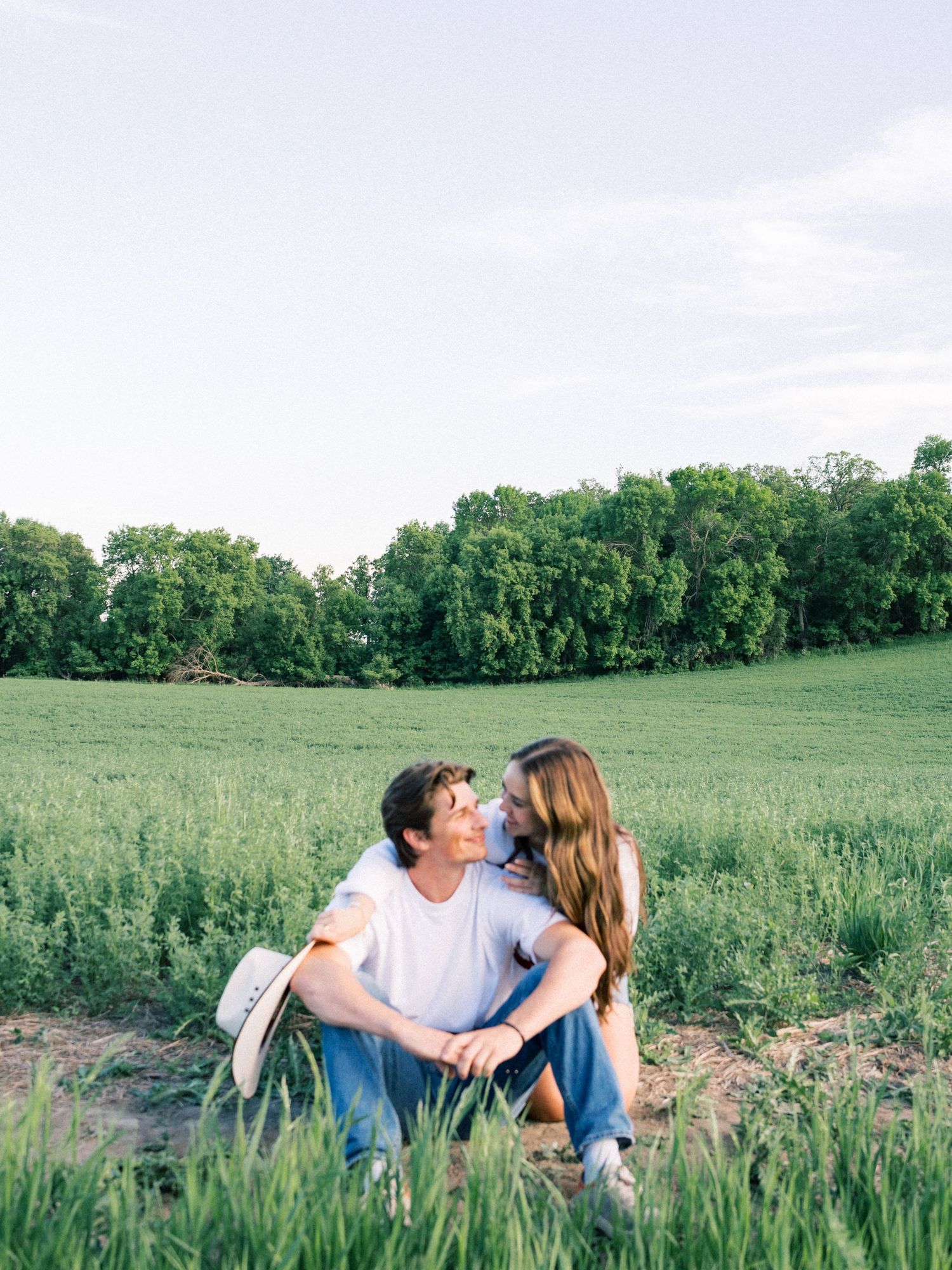 A couple sits together in a lush green field, sharing a tender moment under a soft evening sky.
