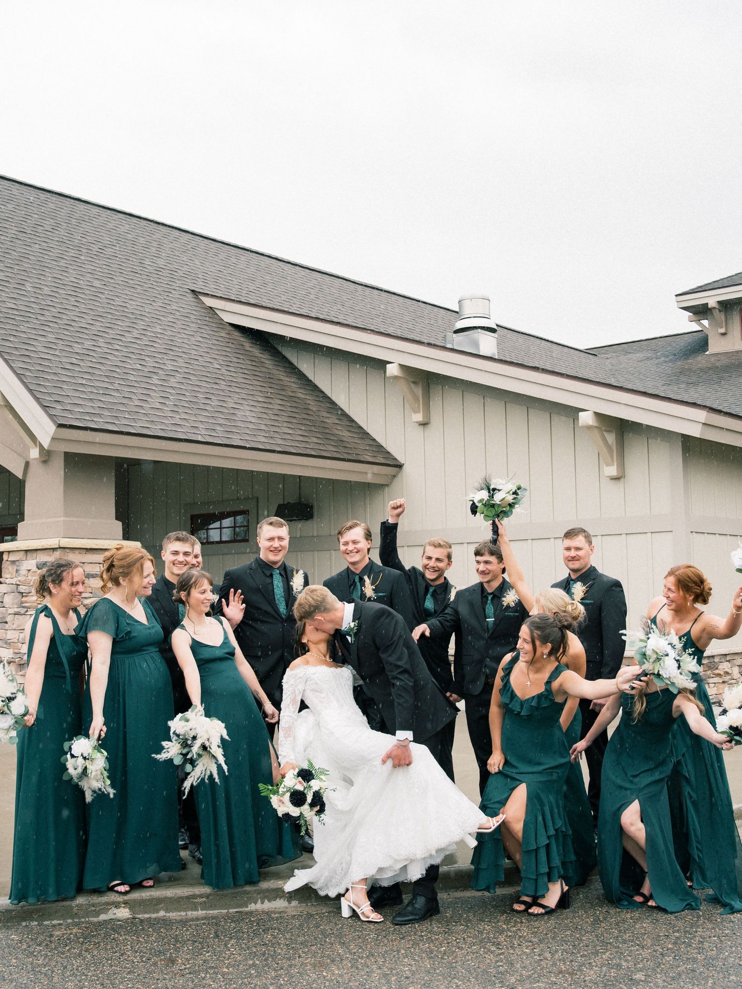 Wedding party in emerald green dresses and black suits celebrating with bride and groom outside rustic venue.