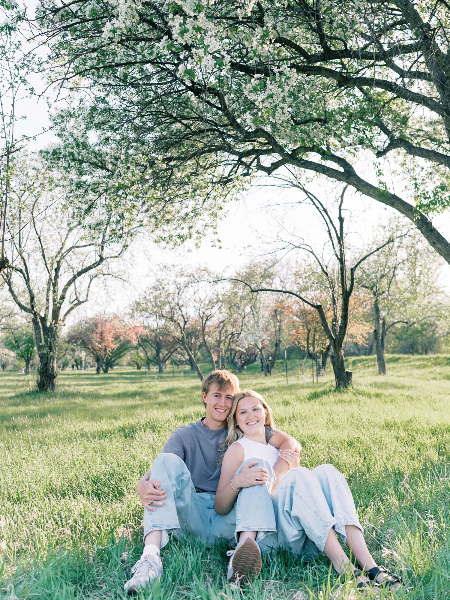 A young couple sits together on green grass under blooming spring trees, smiling warmly in casual light blue outfits.
