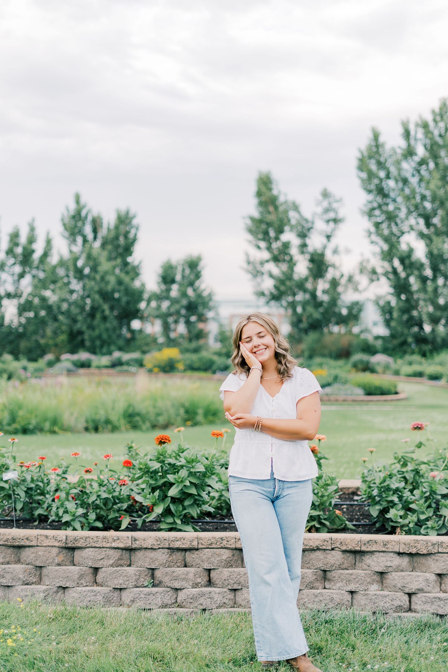 Smiling person stands in a garden with red flowers and a stone retaining wall on a cloudy summer day.
