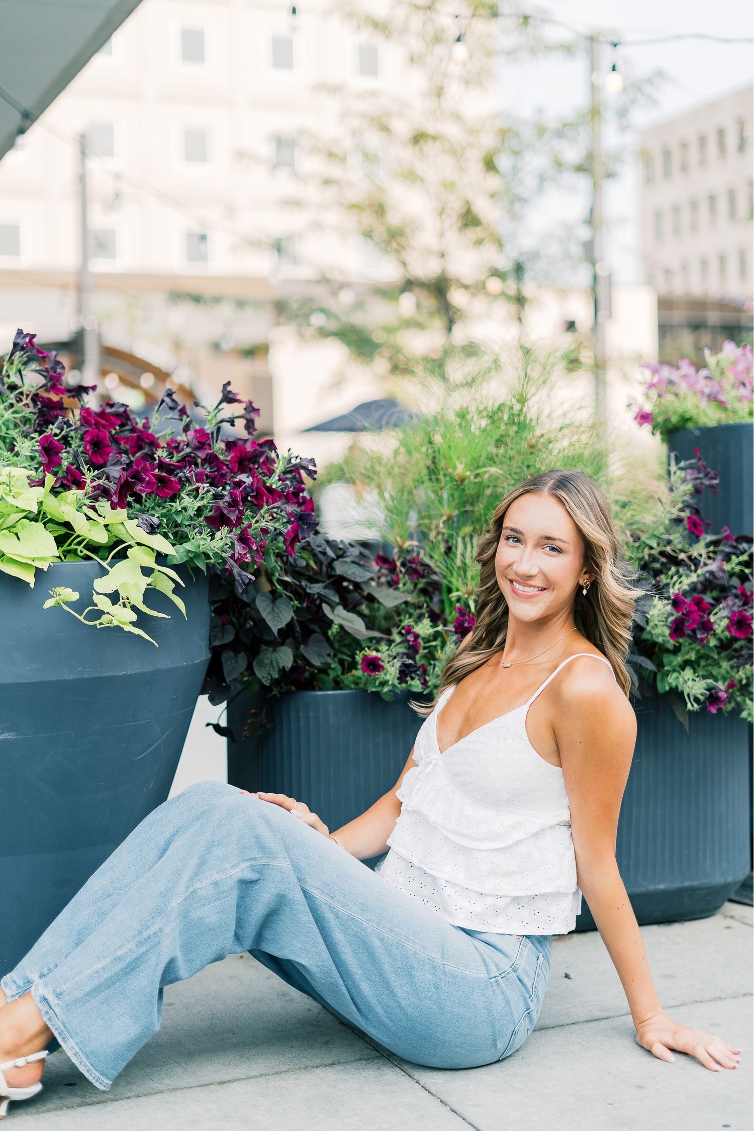 A person in a white tank top and jeans sits casually by decorative planters filled with purple flowers on a city sidewalk.