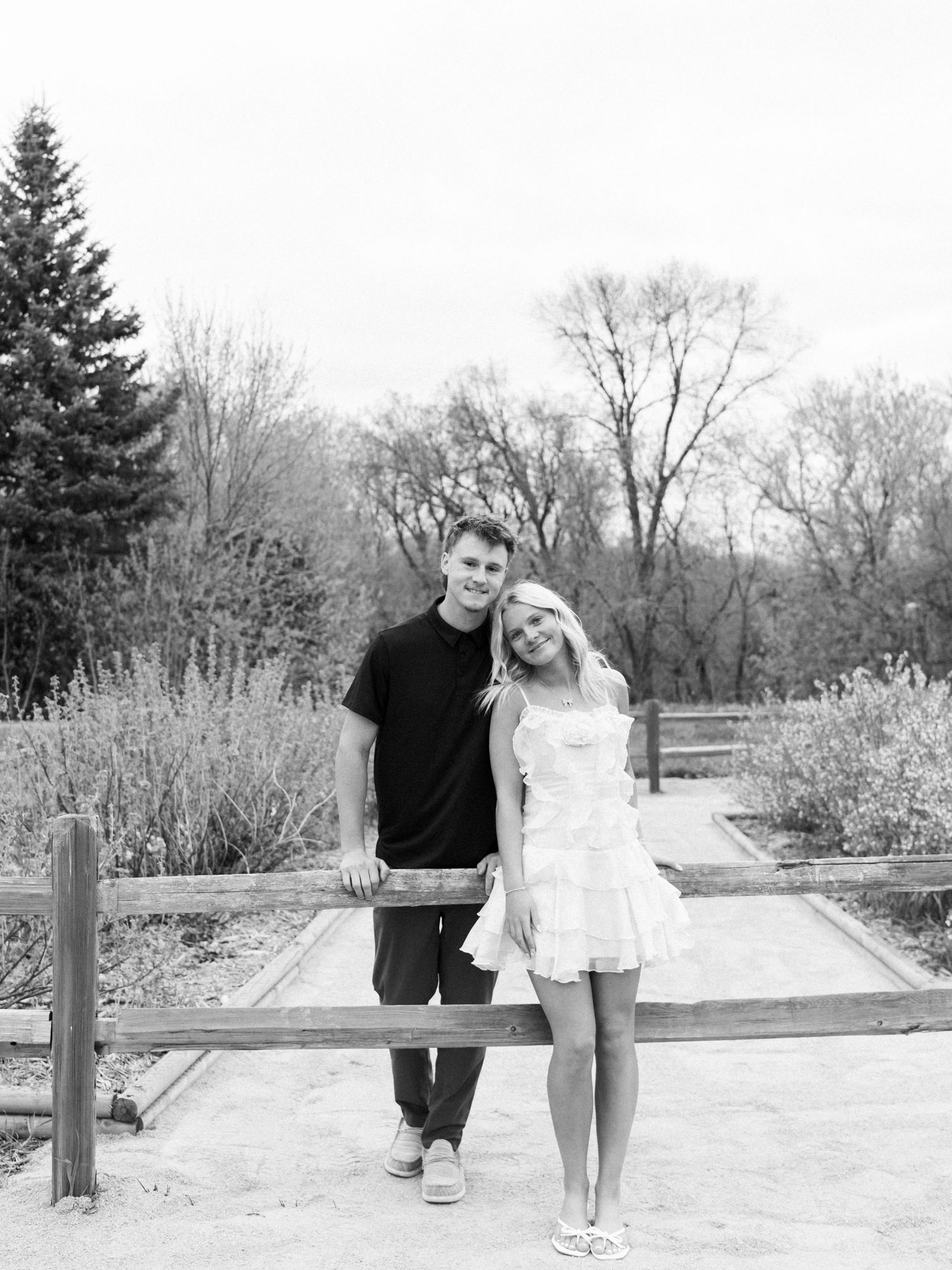 Black and white photo of a young couple smiling together by a wooden fence in a park setting.