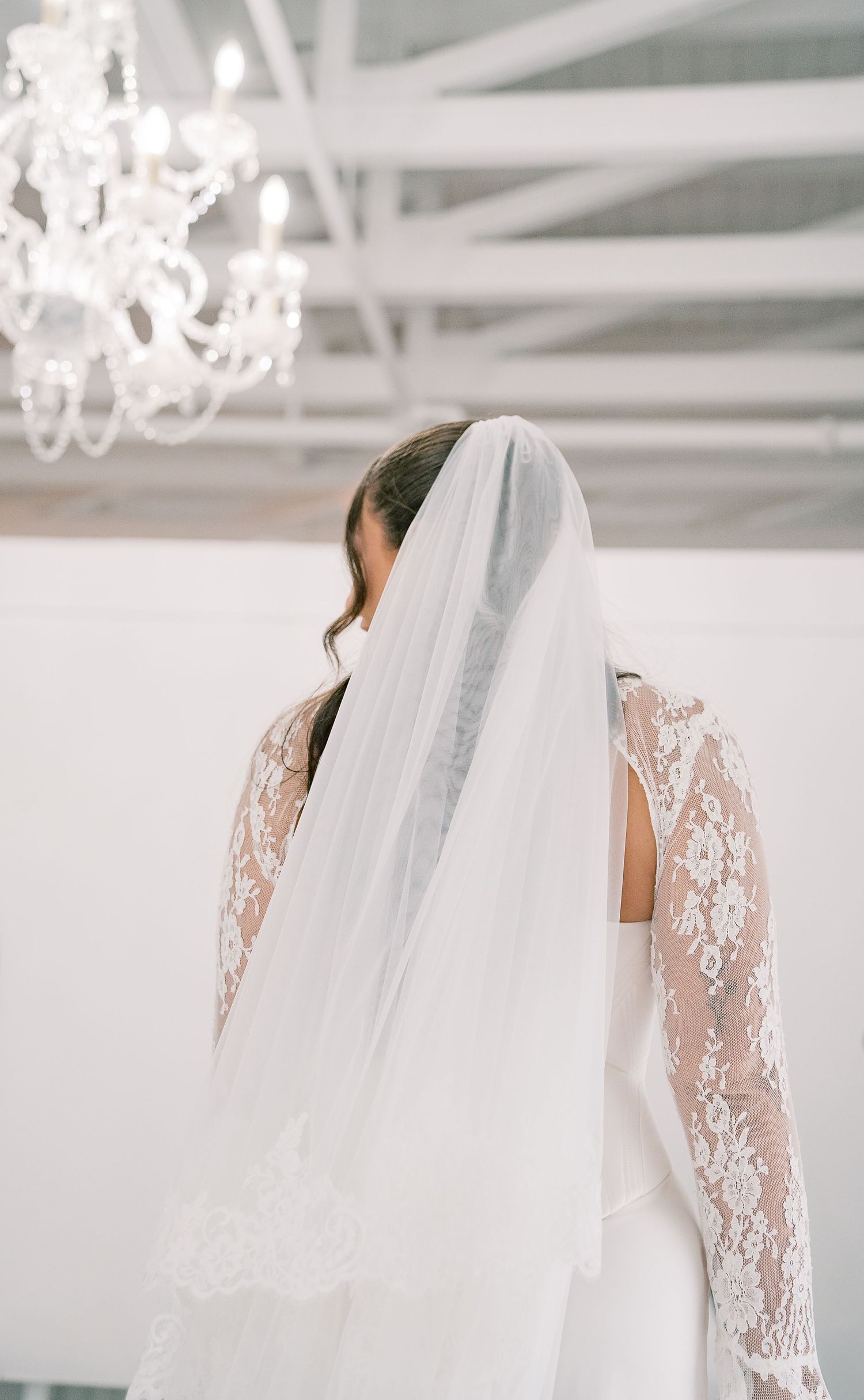 A bride in a delicate lace sleeve wedding gown with flowing veil stands in an ethereal white setting.