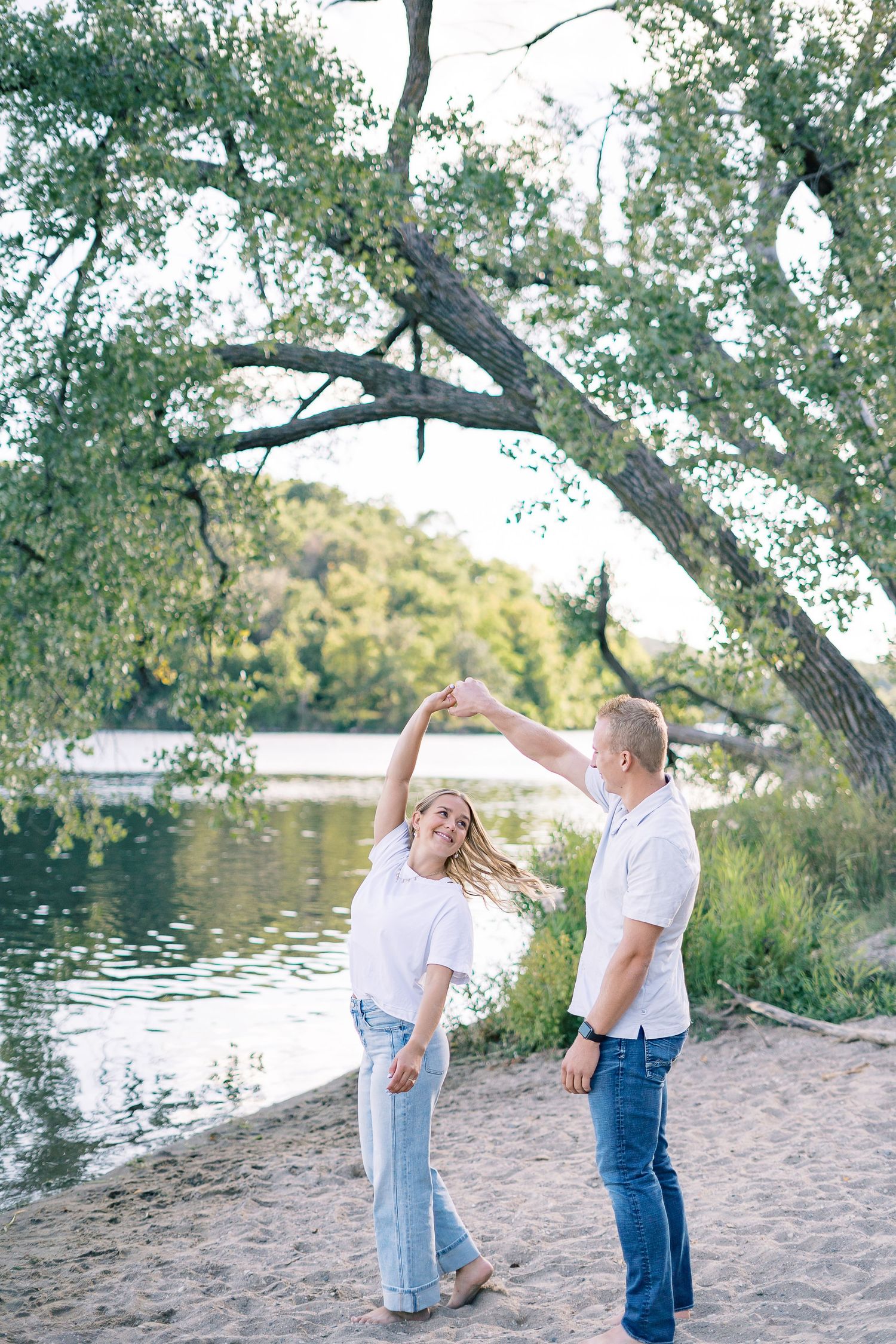 A romantic couple embraces on a sandy beach near a lake with overhanging trees in the background.