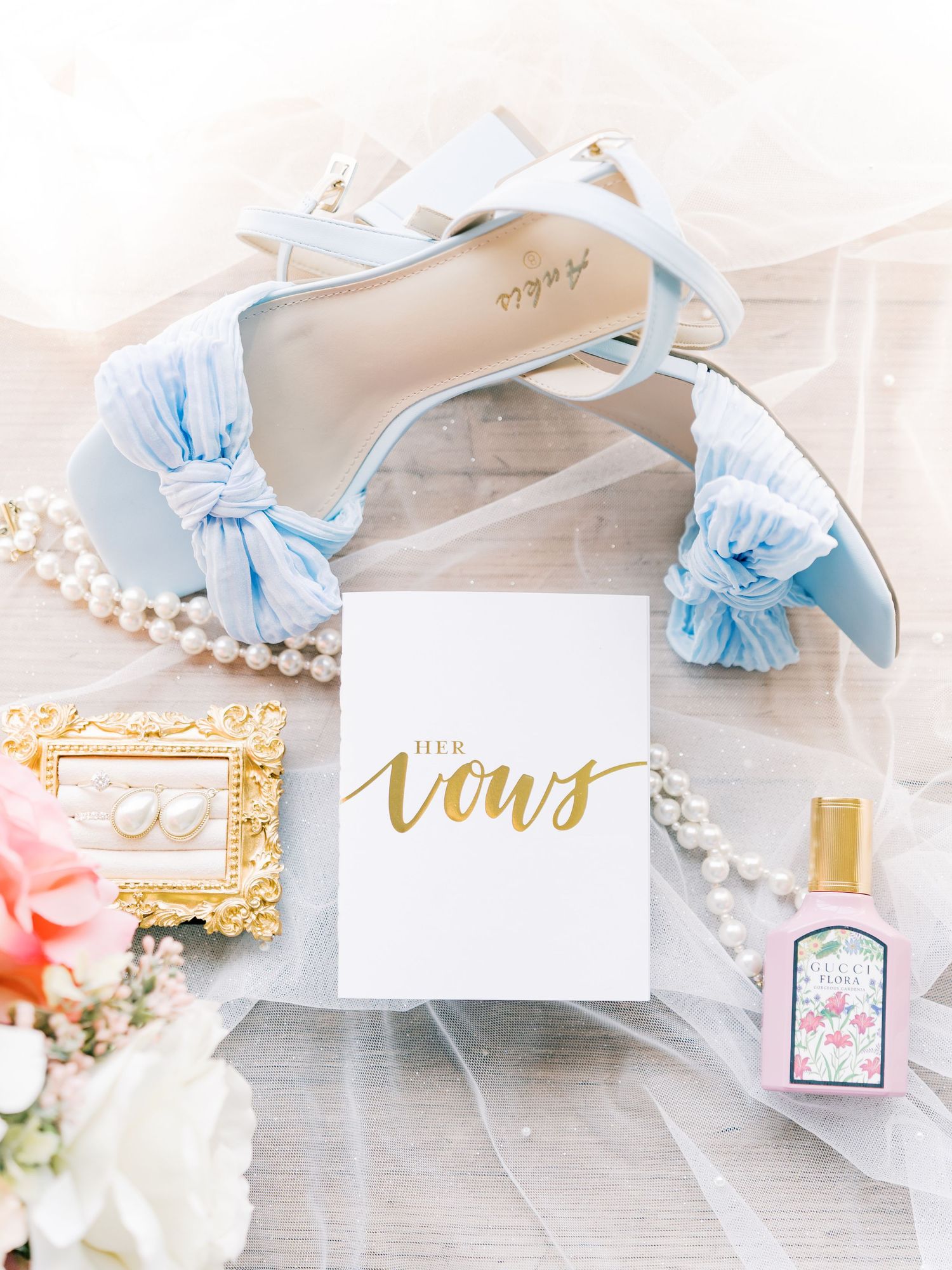 Light blue bridal heels with bow detail beside vows book, pearl necklace, floral earrings, and Gucci perfume on soft fabric.