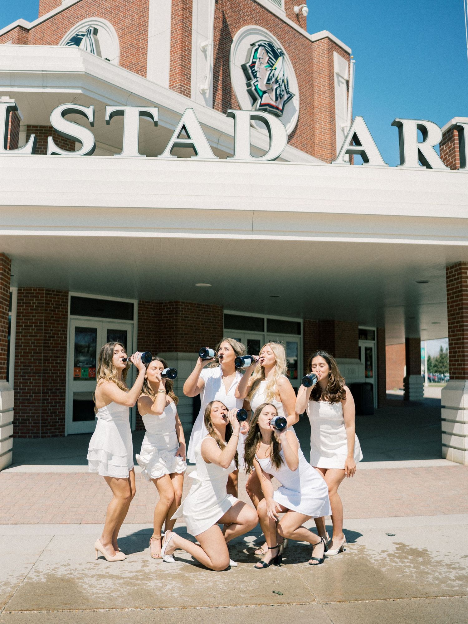 Group of women in white dresses posing and drinking champagne outside Engelstad Arena on a sunny day.
