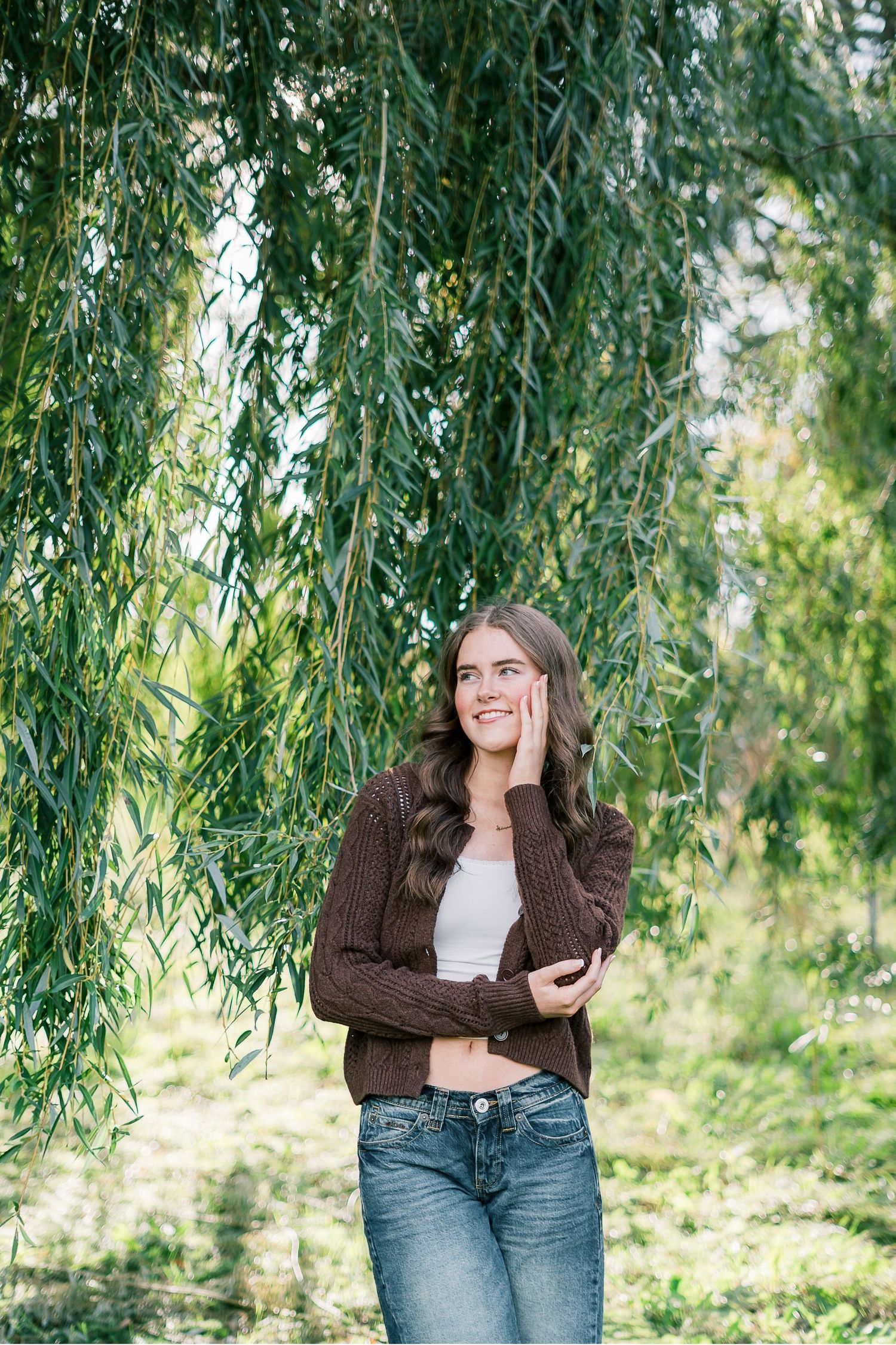 Person in brown sweater and jeans poses beneath cascading willow tree branches in outdoor setting.