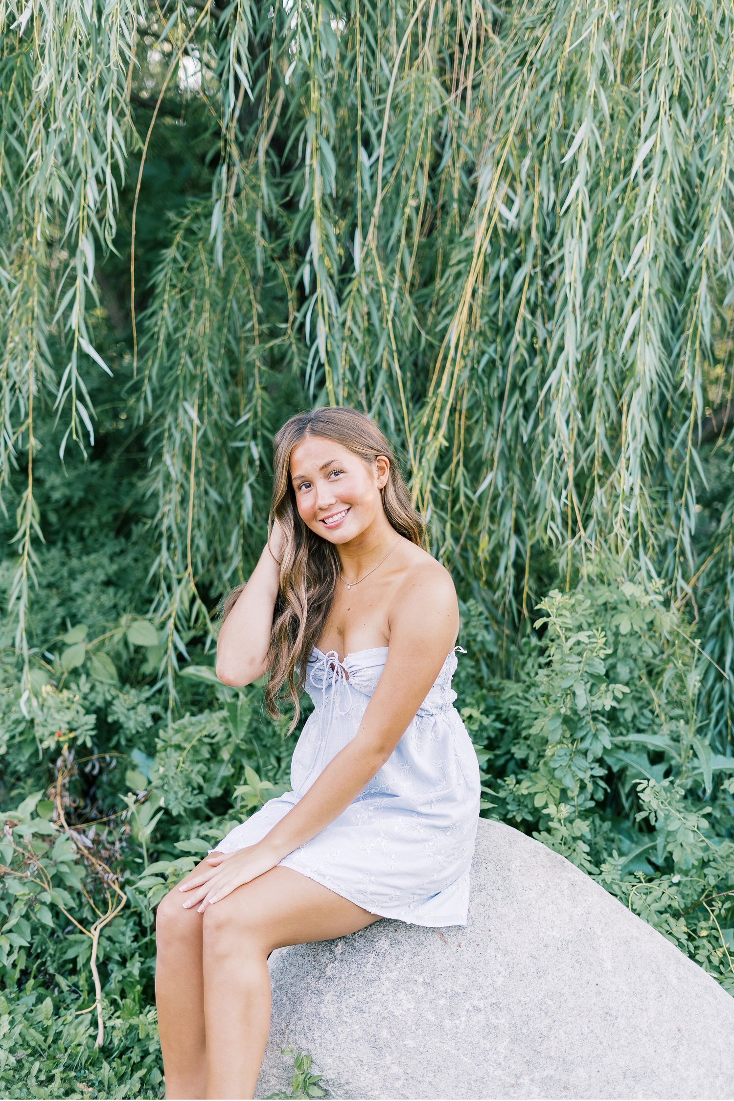 A person in a white dress poses outdoors on a rock under drooping willow tree branches.