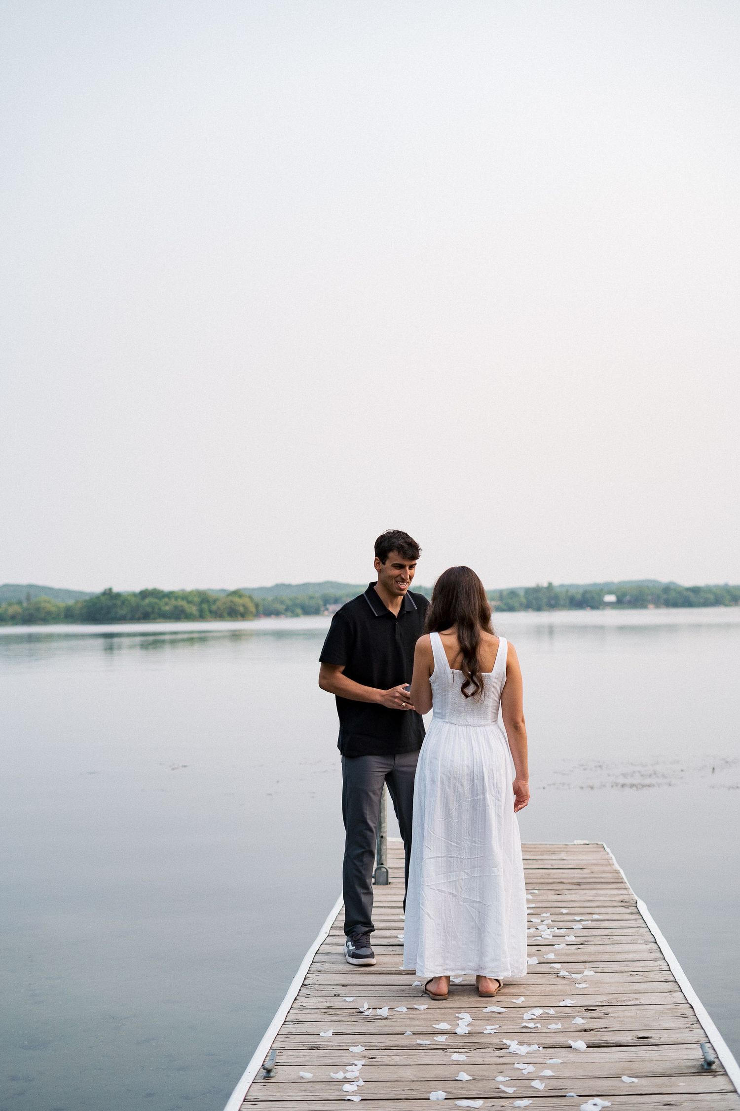 A couple walks together on a wooden dock overlooking a serene lake on a misty day.