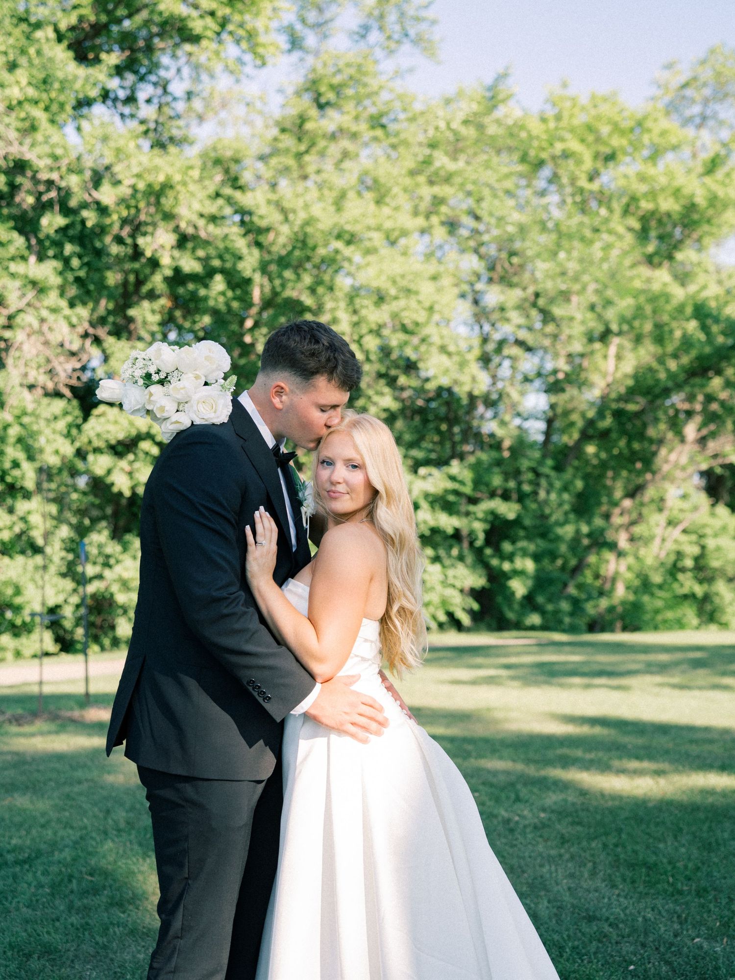 A bride and groom embrace outdoors, she holds white flowers in a satin gown as he kisses her head in a black tuxedo.