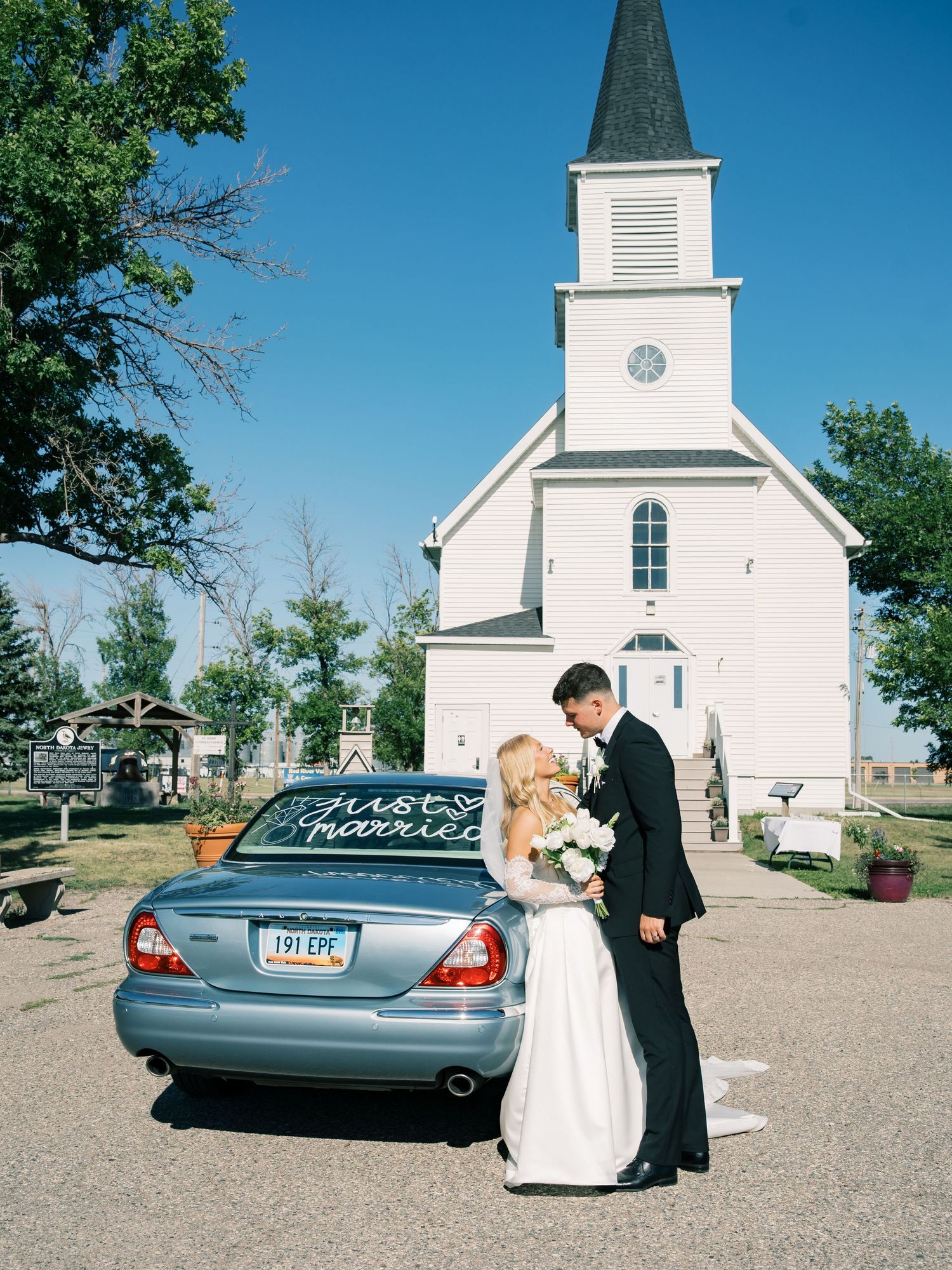 Newlywed couple kissing beside a Just Married car in front of a white church on their wedding day.
