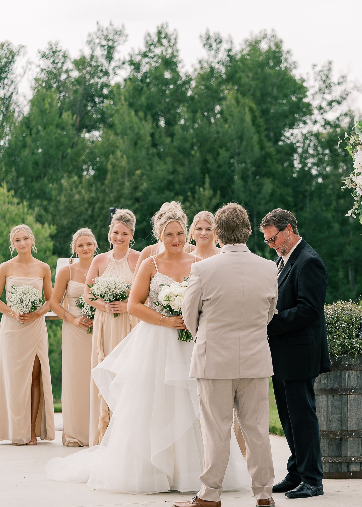 An outdoor wedding ceremony taking place on a patio with bridesmaids in beige dresses and natural greenery in background.