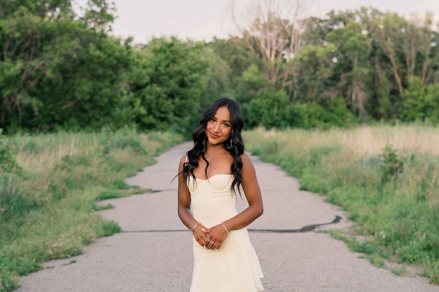 A person in a white dress poses outdoors in a natural setting with trees and grass along a pathway.