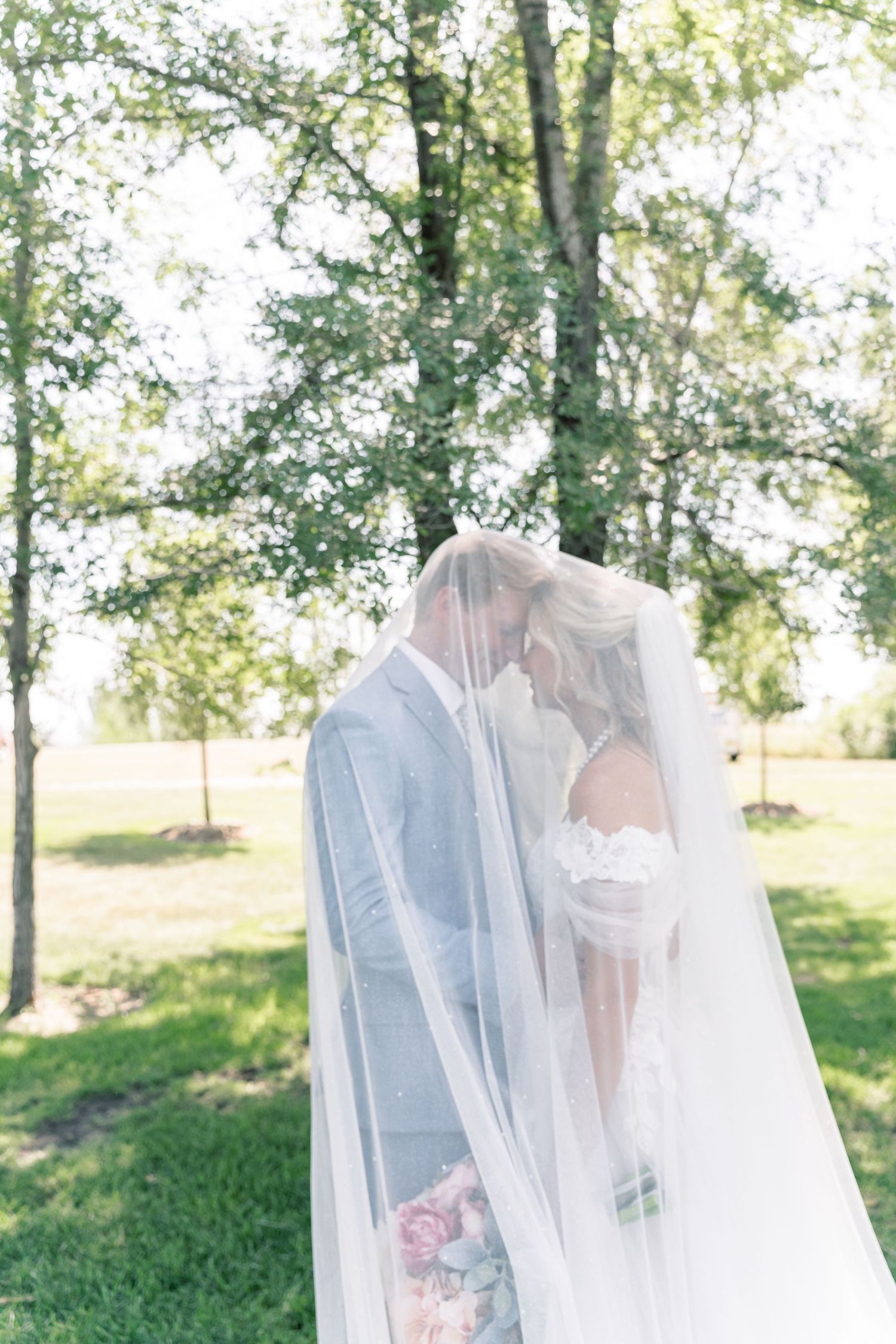 Bride and groom sharing a romantic kiss under a flowing white veil outdoors surrounded by lush green trees.