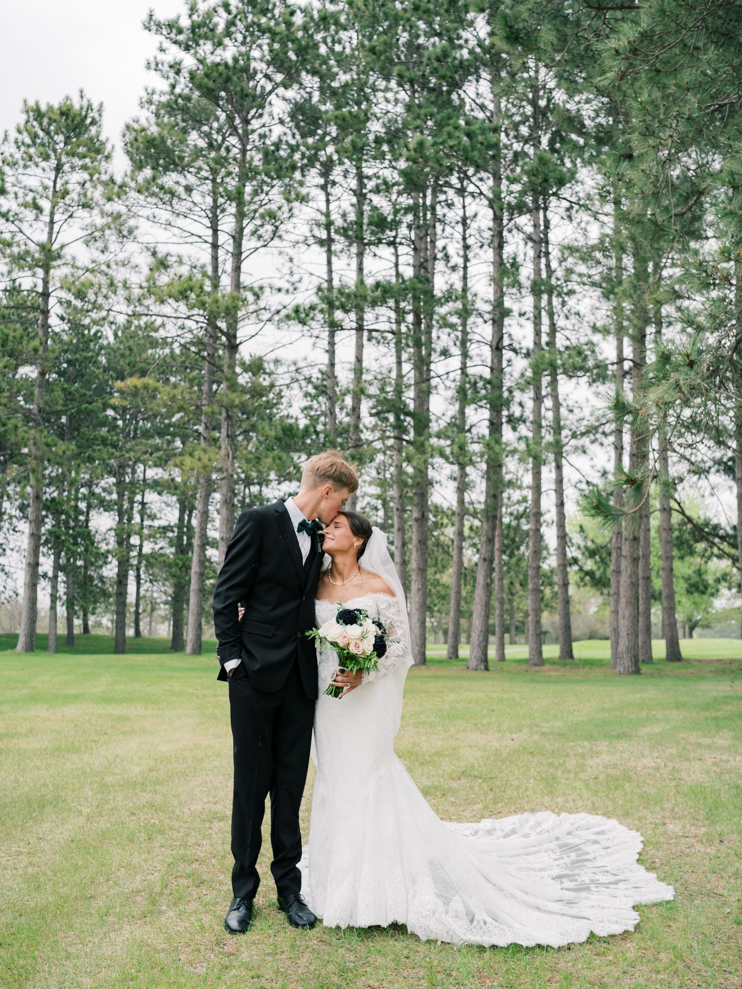 A bride and groom share a romantic moment in a lush green field surrounded by tall pine trees on their wedding day.