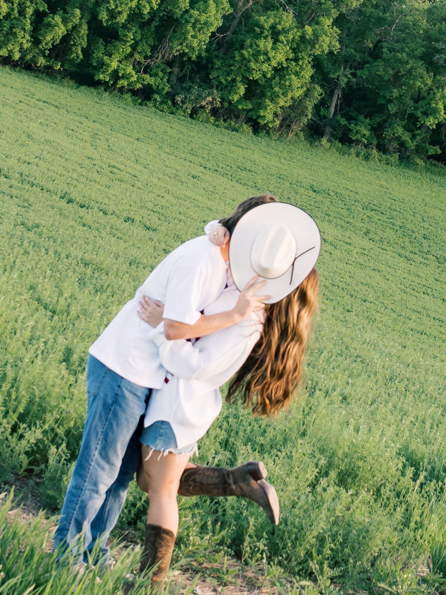 Couple sharing a romantic kiss in a green field, woman wearing cowboy hat and brown boots with long flowing hair.