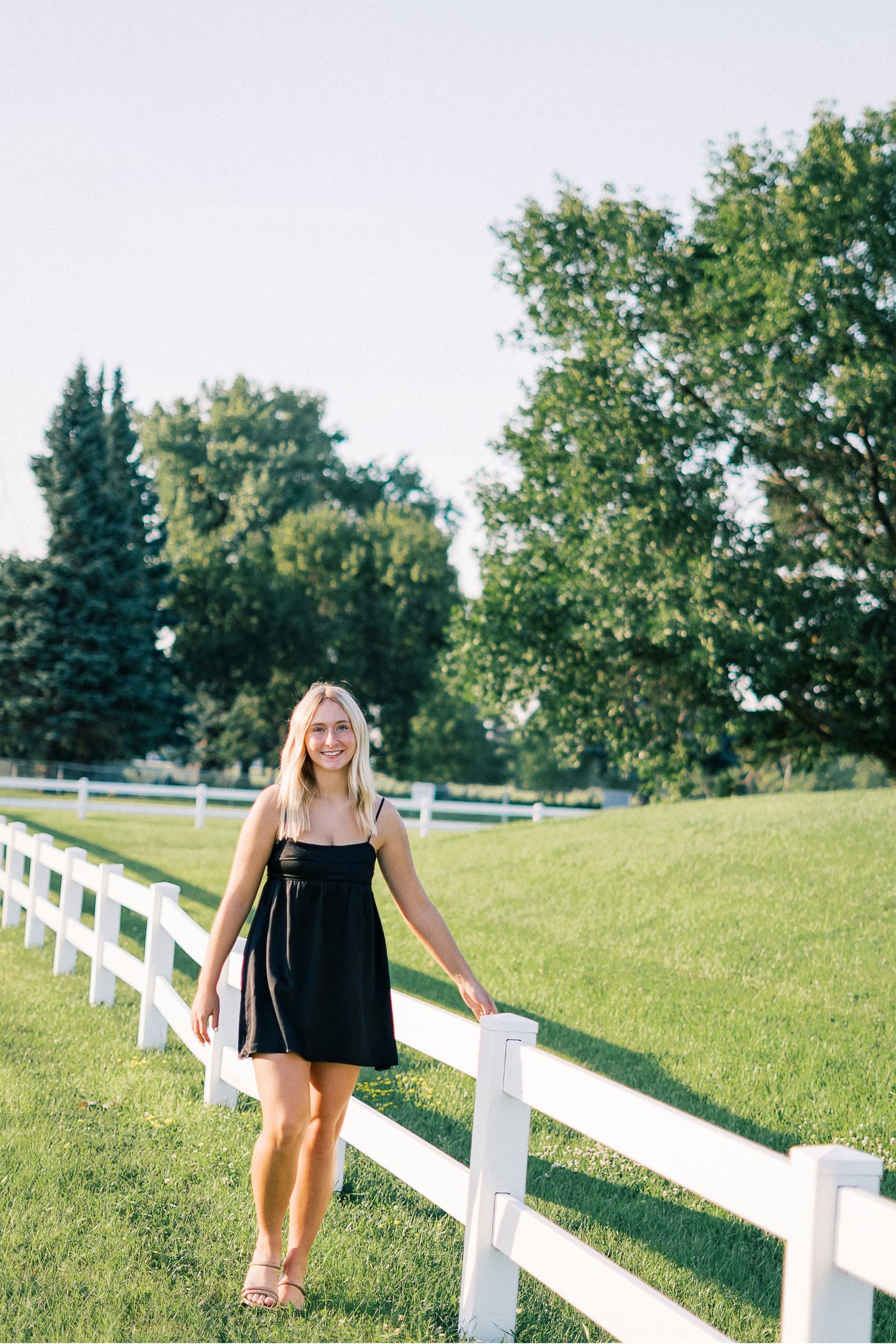 A person in a black sundress stands along a white wooden fence on a sunny summer day with green trees in the background.