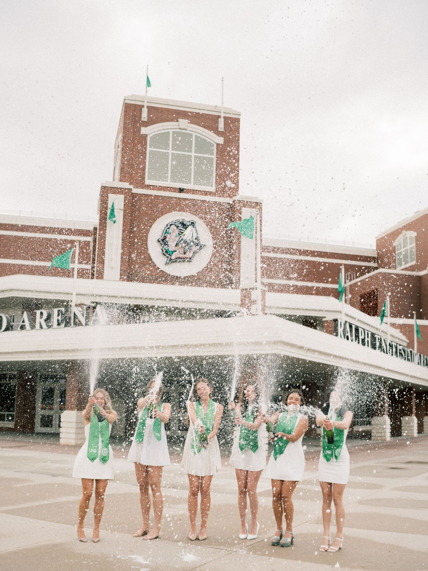 Six cheerleaders in white and green uniforms celebrate with confetti in front of a brick arena building.
