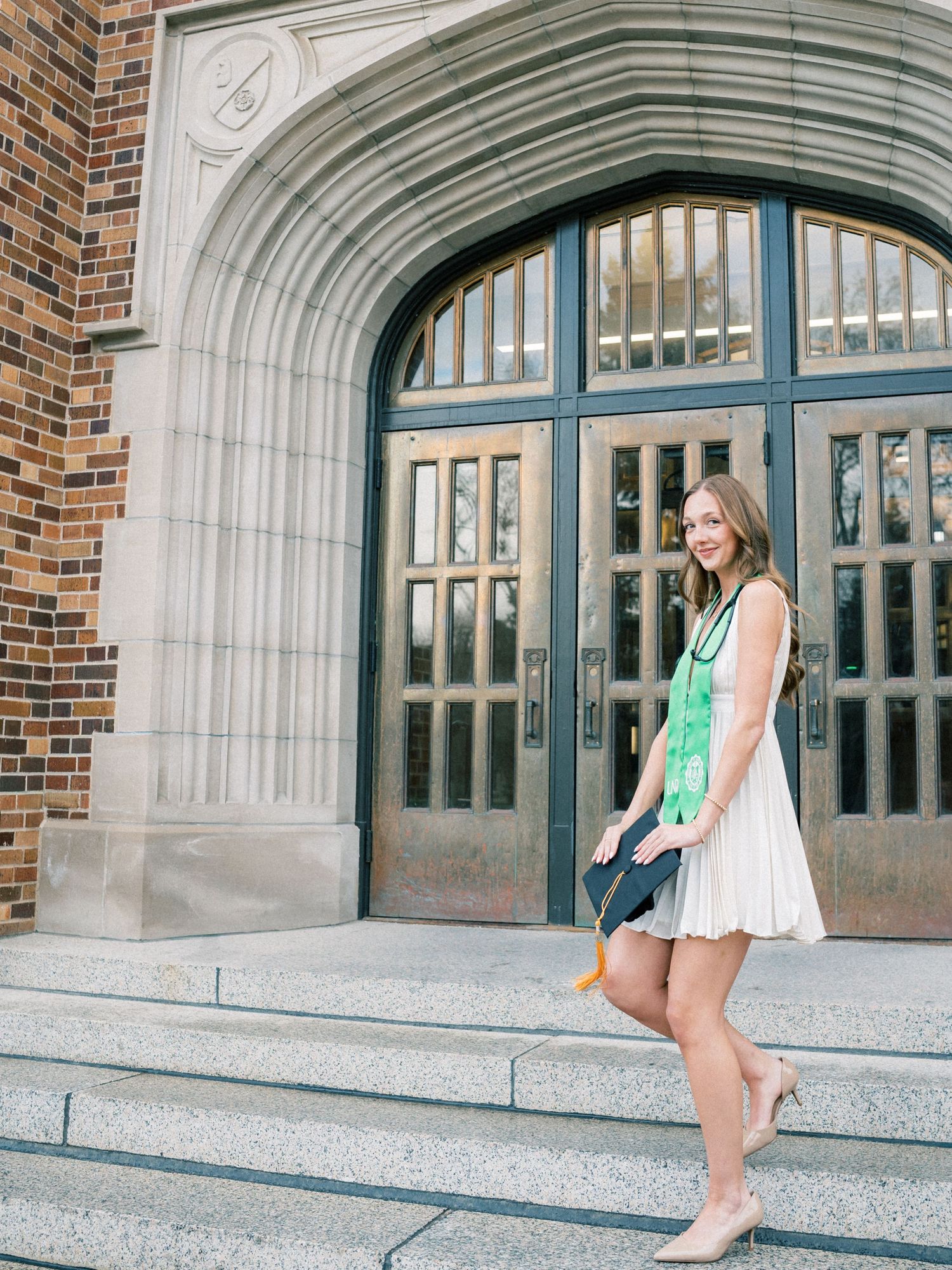 Woman in white dress with green graduation sash poses on university building steps.