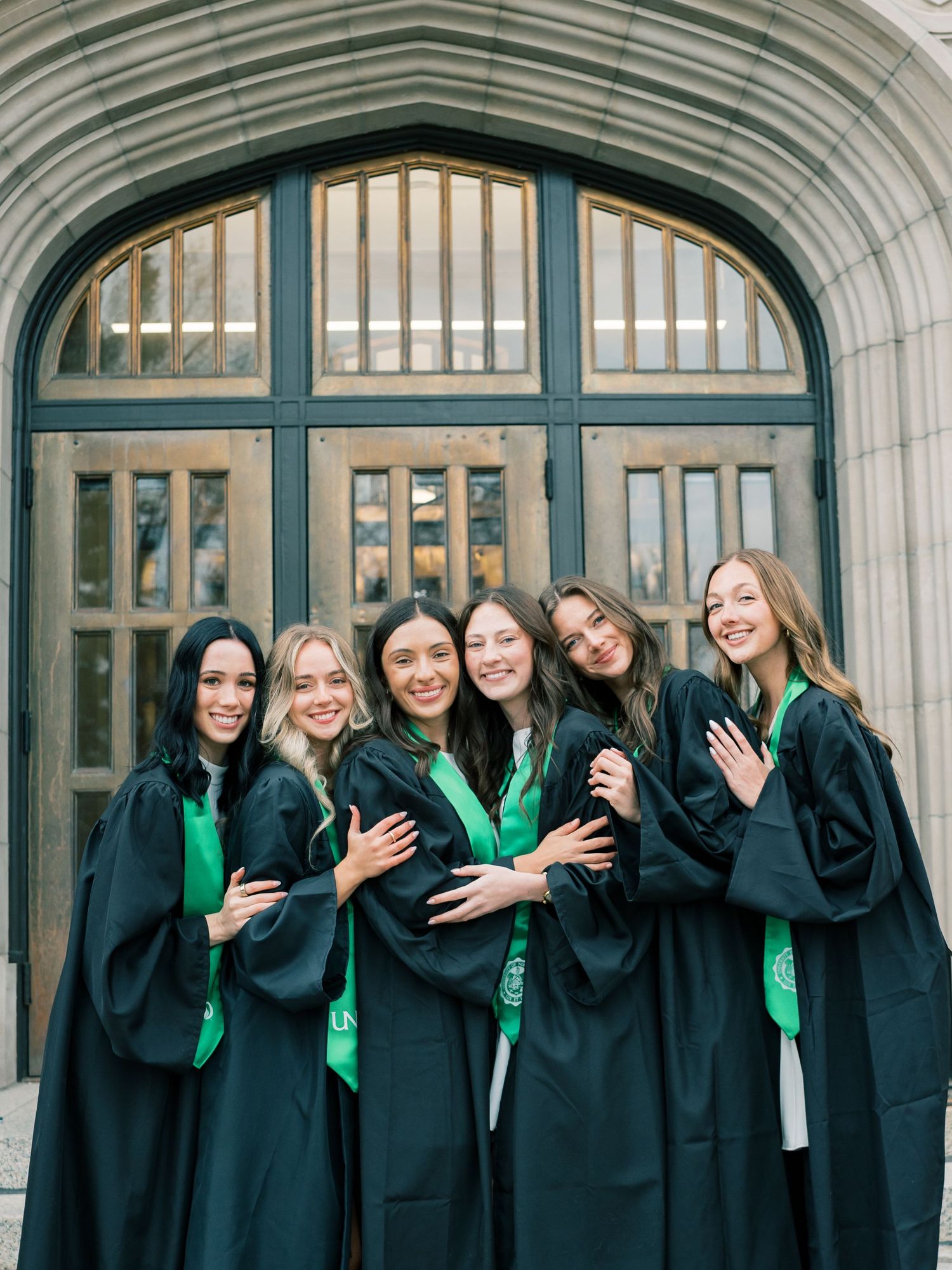 Six women in black and green graduation gowns pose together smiling in front of an arched stone building entrance.