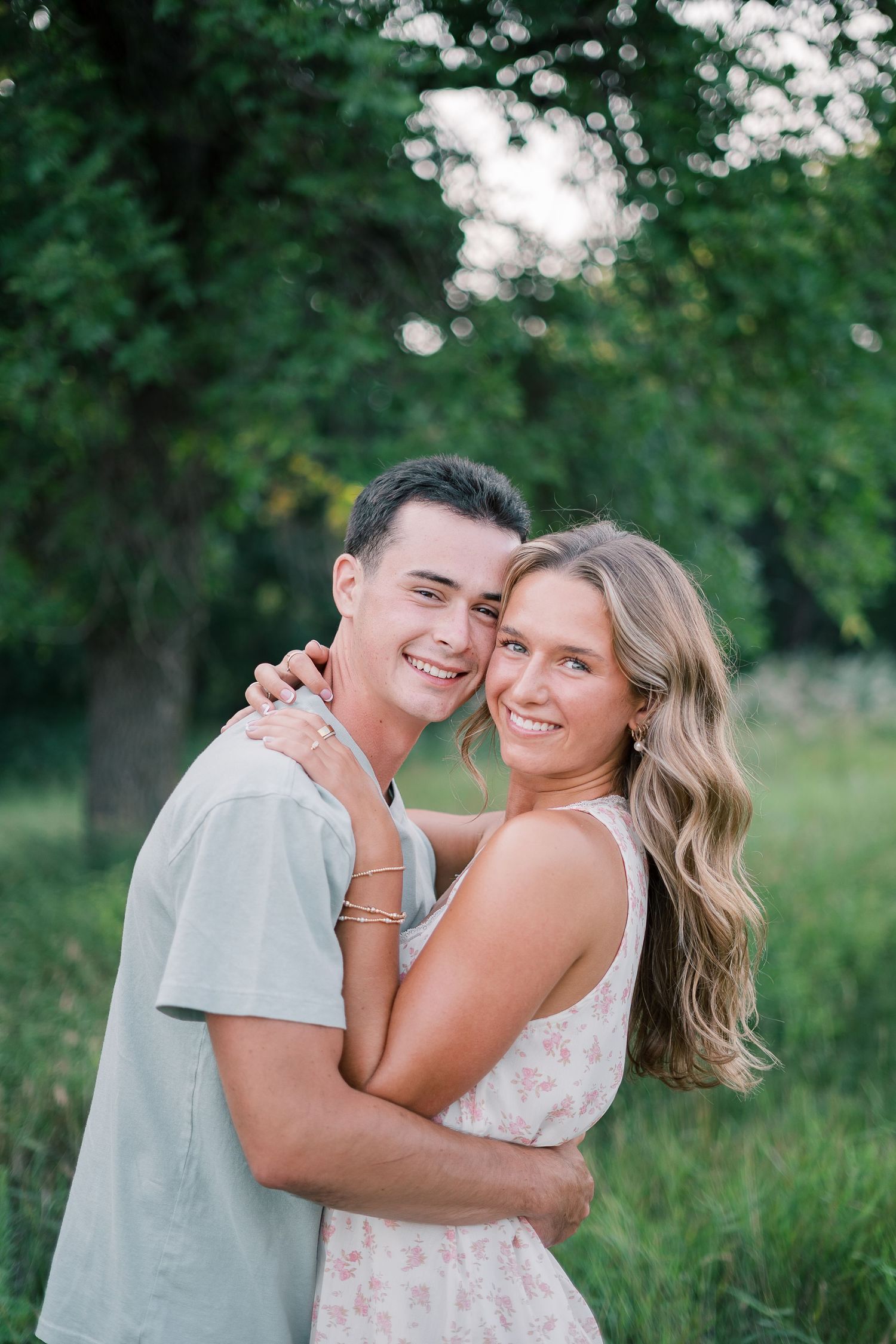 A couple embraces and smiles together outdoors with lush green foliage in the background.