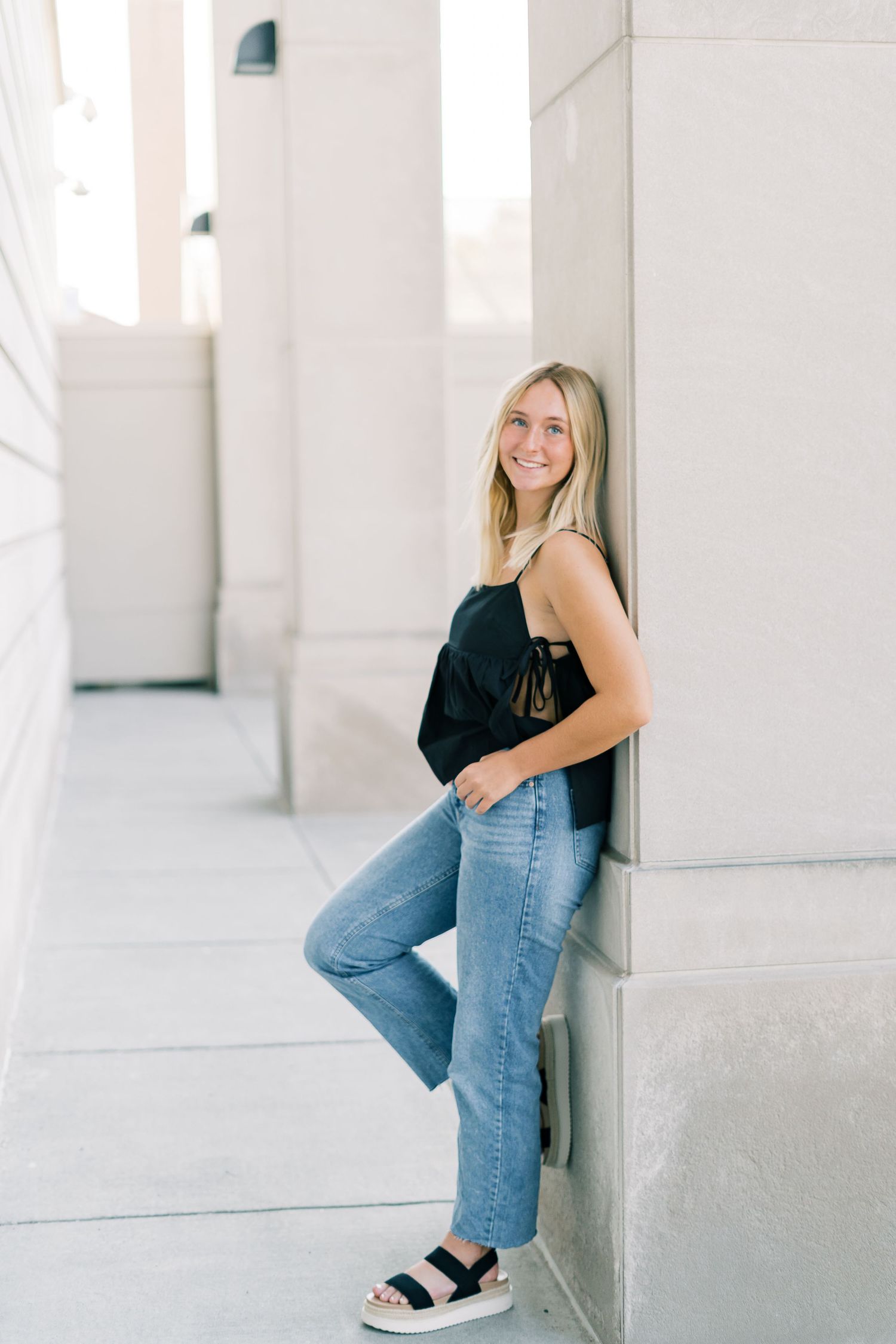Young woman in jeans and black top poses casually against white interior walls.