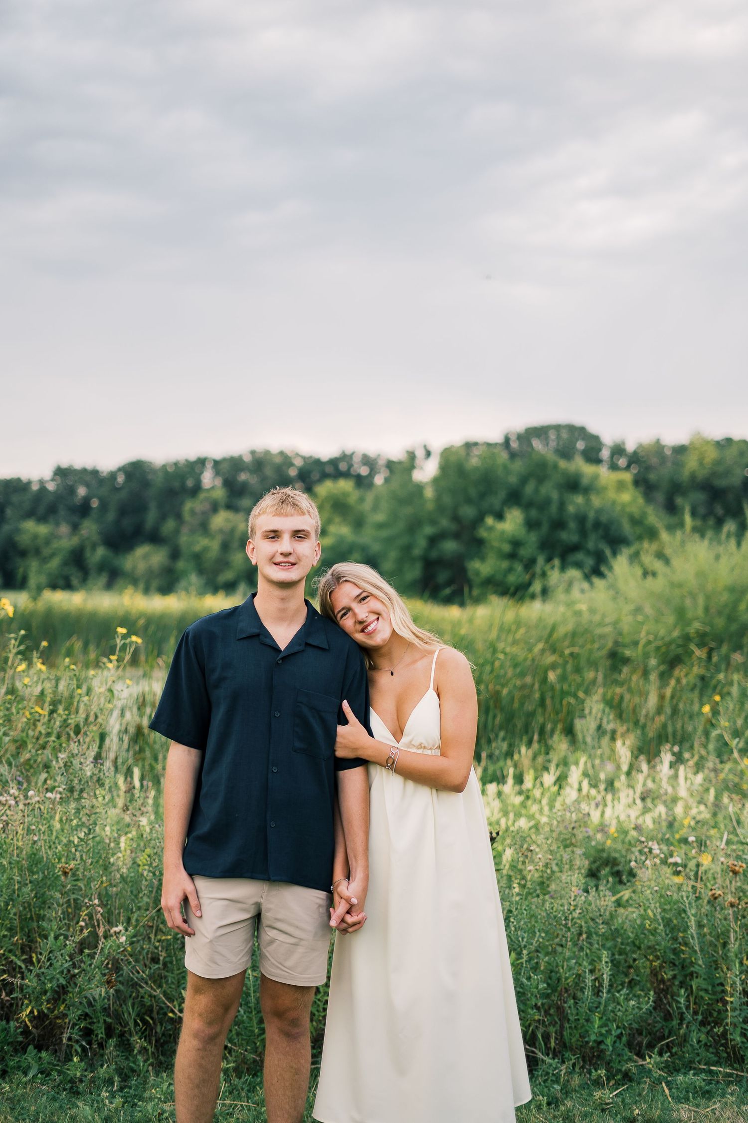 A couple posing together in a serene meadow with tall grass and trees in the background on a sunny day.