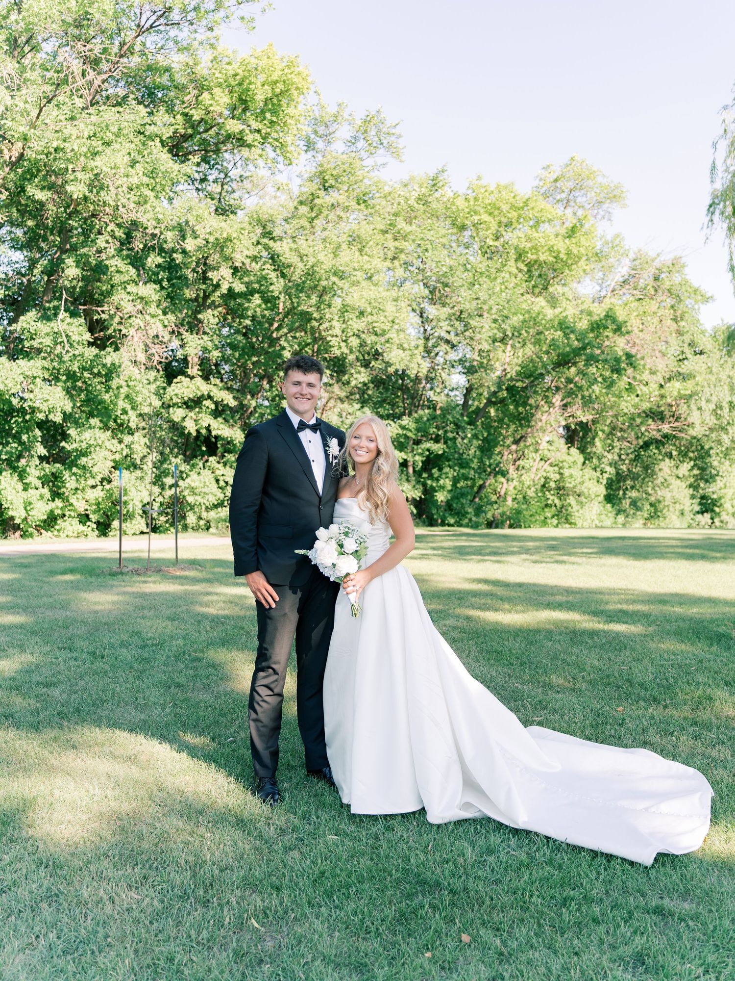Bride and groom posing on green lawn in wedding attire, bride holding white bouquet, lush trees in background.