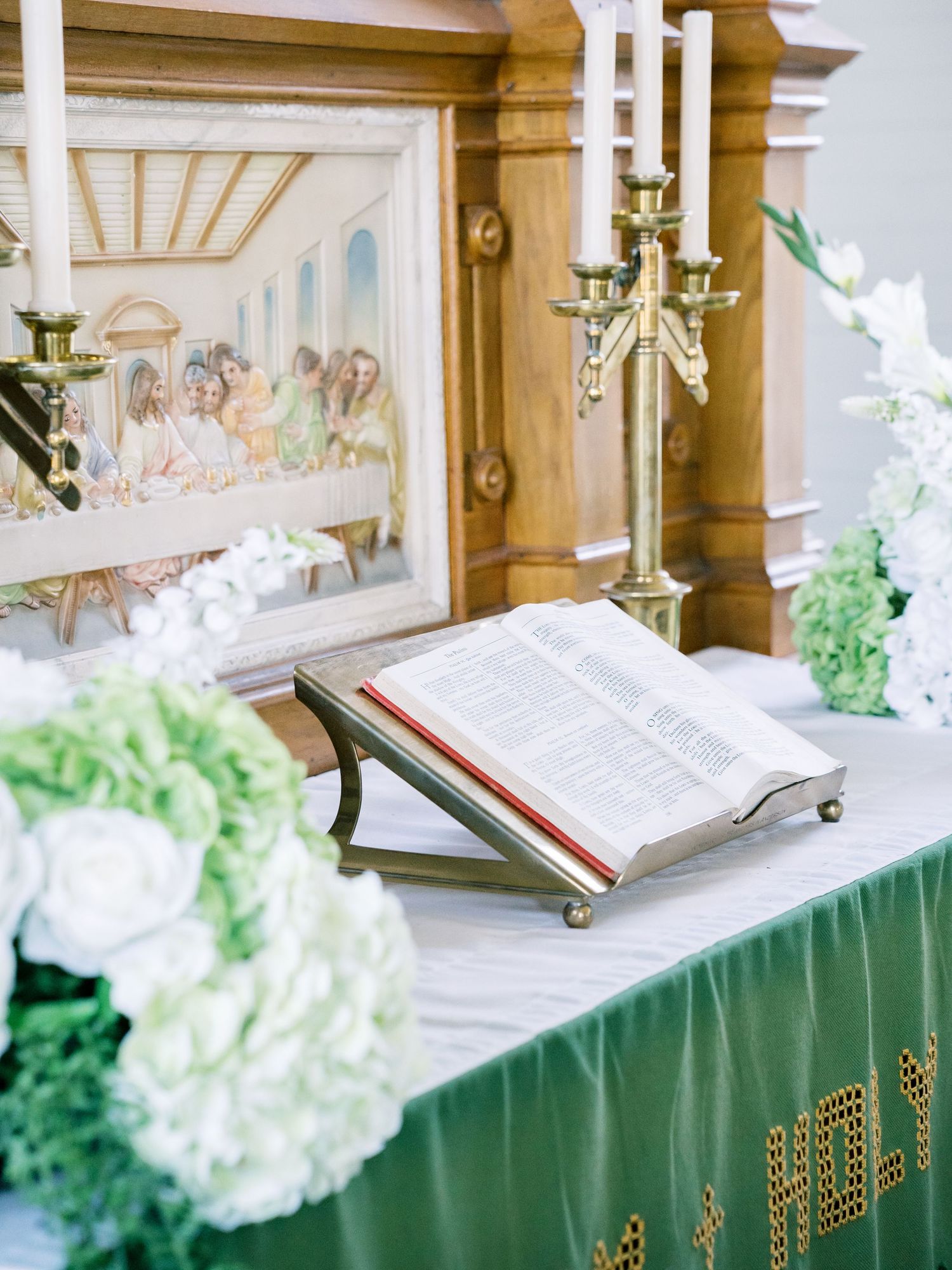 Open Bible on brass stand beside green floral arrangements and Last Supper painting at elegant wedding altar.