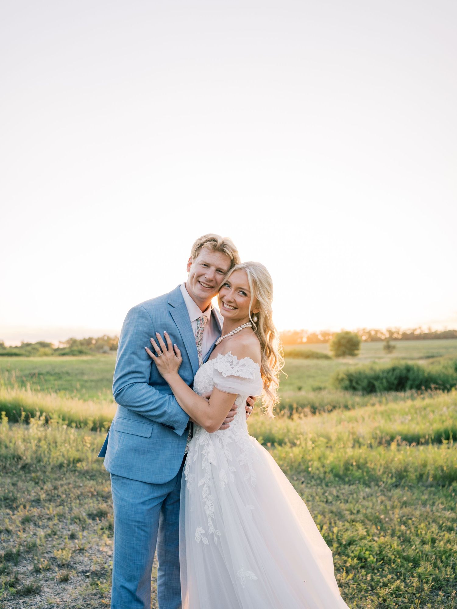 A couple poses in a golden field at sunset, the groom in a blue suit and bride in an off-shoulder white gown.