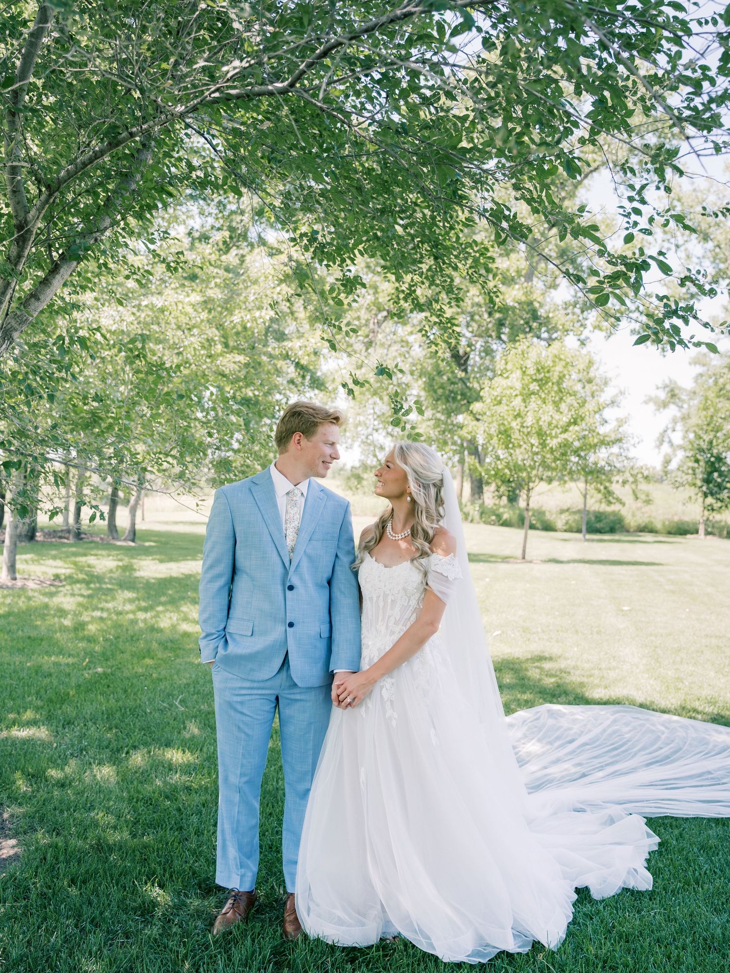 Bride in flowing white gown and groom in light blue suit holding hands in lush green garden setting.