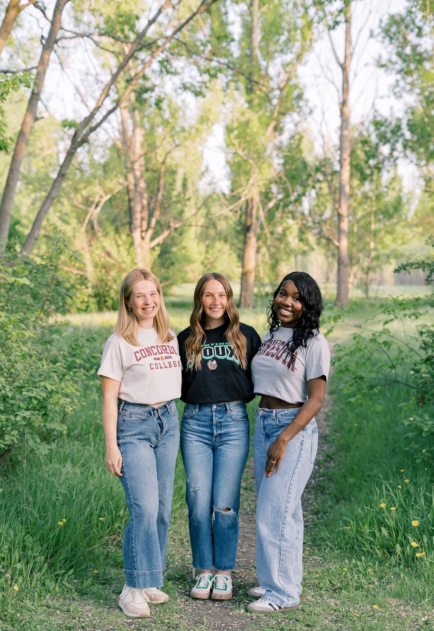 Three friends wearing casual college t-shirts and jeans pose together on a dirt path surrounded by green trees and grass.