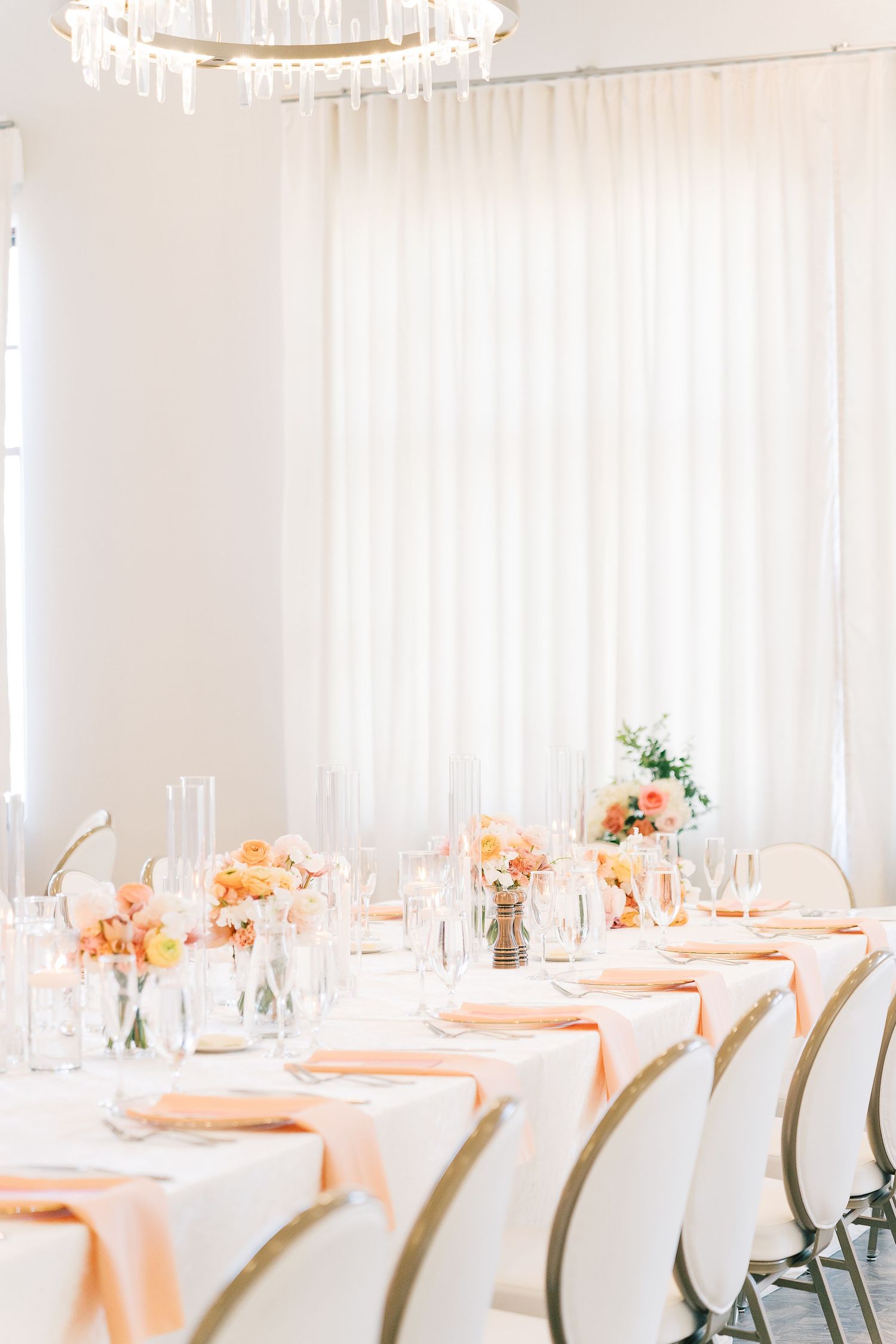 Elegant wedding reception tables set with peach linens and white chairs in a bright, airy venue with natural lighting.