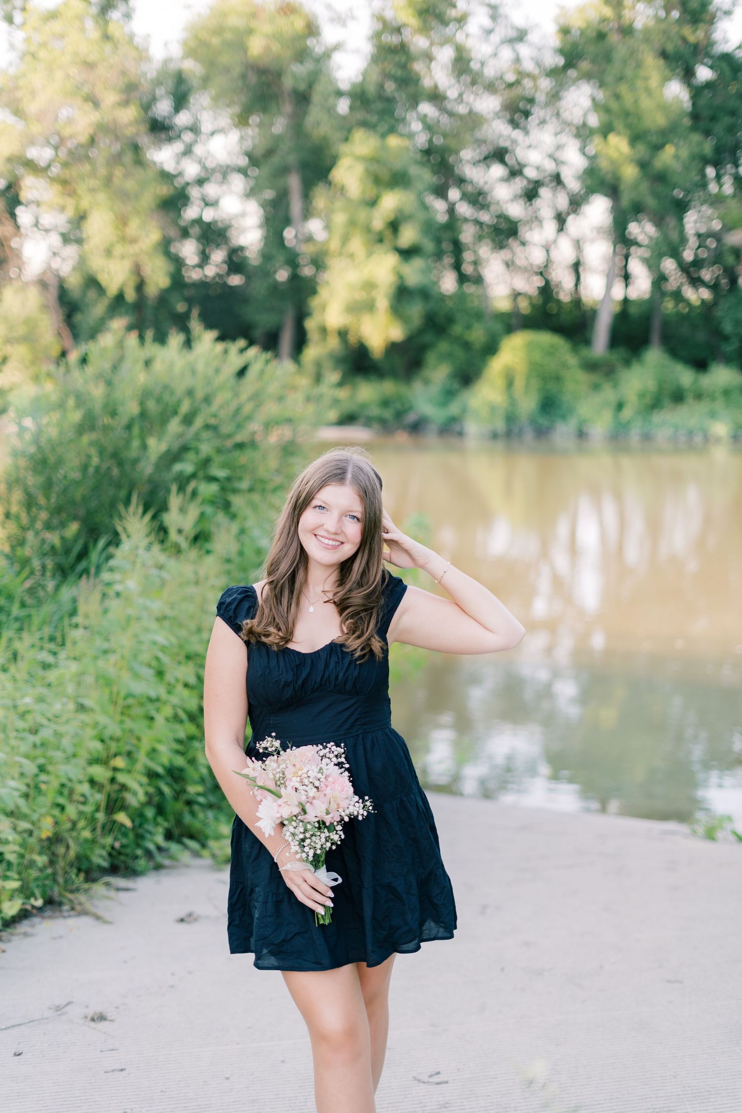 Someone in a navy blue dress holds dried flowers while posing on a path near a serene lake surrounded by greenery.