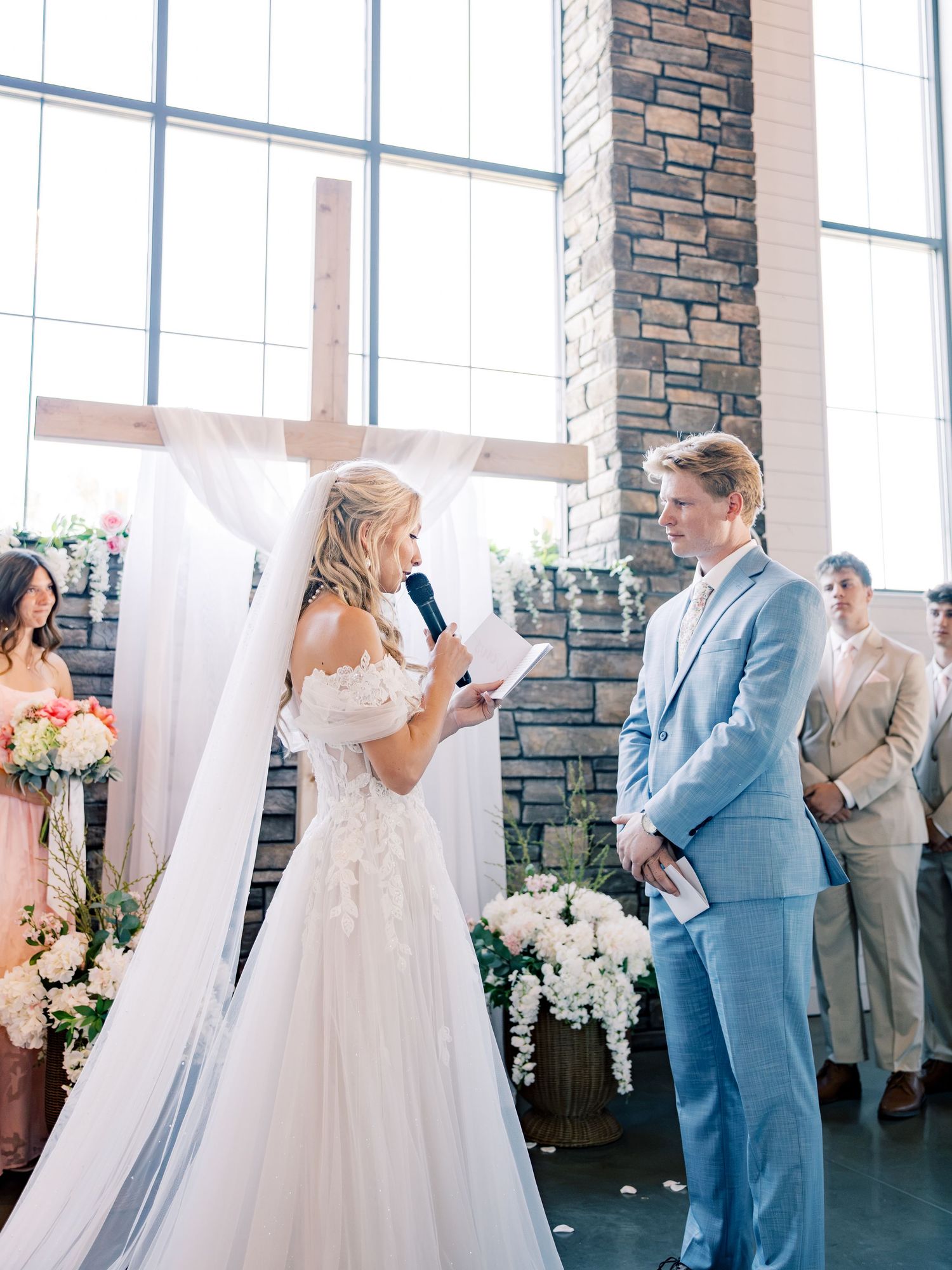 Bride reads vows to groom at elegant church wedding ceremony with wooden cross, floral arrangements, and bridal party.