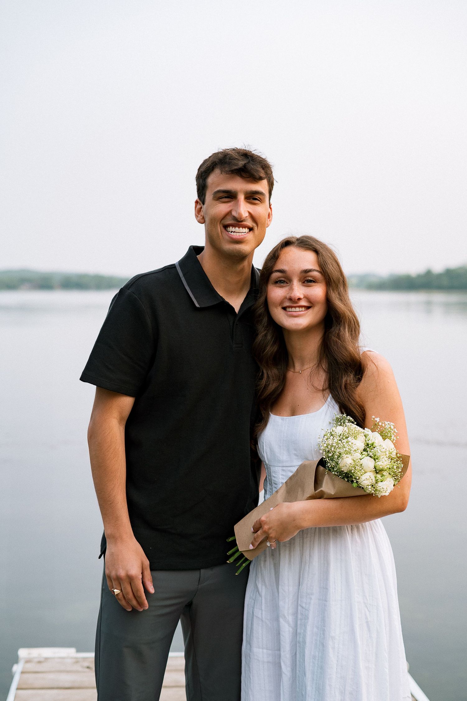 A couple poses together by a waterfront, with the woman wearing a white dress and holding a small bouquet.