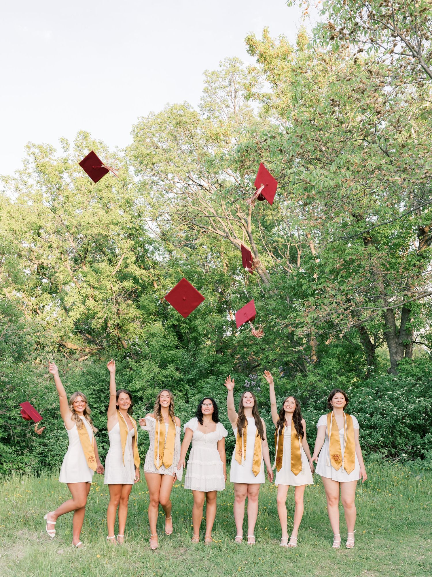Eight female graduates in white dresses toss red mortarboard caps in the air outdoors, celebrating their graduation day.