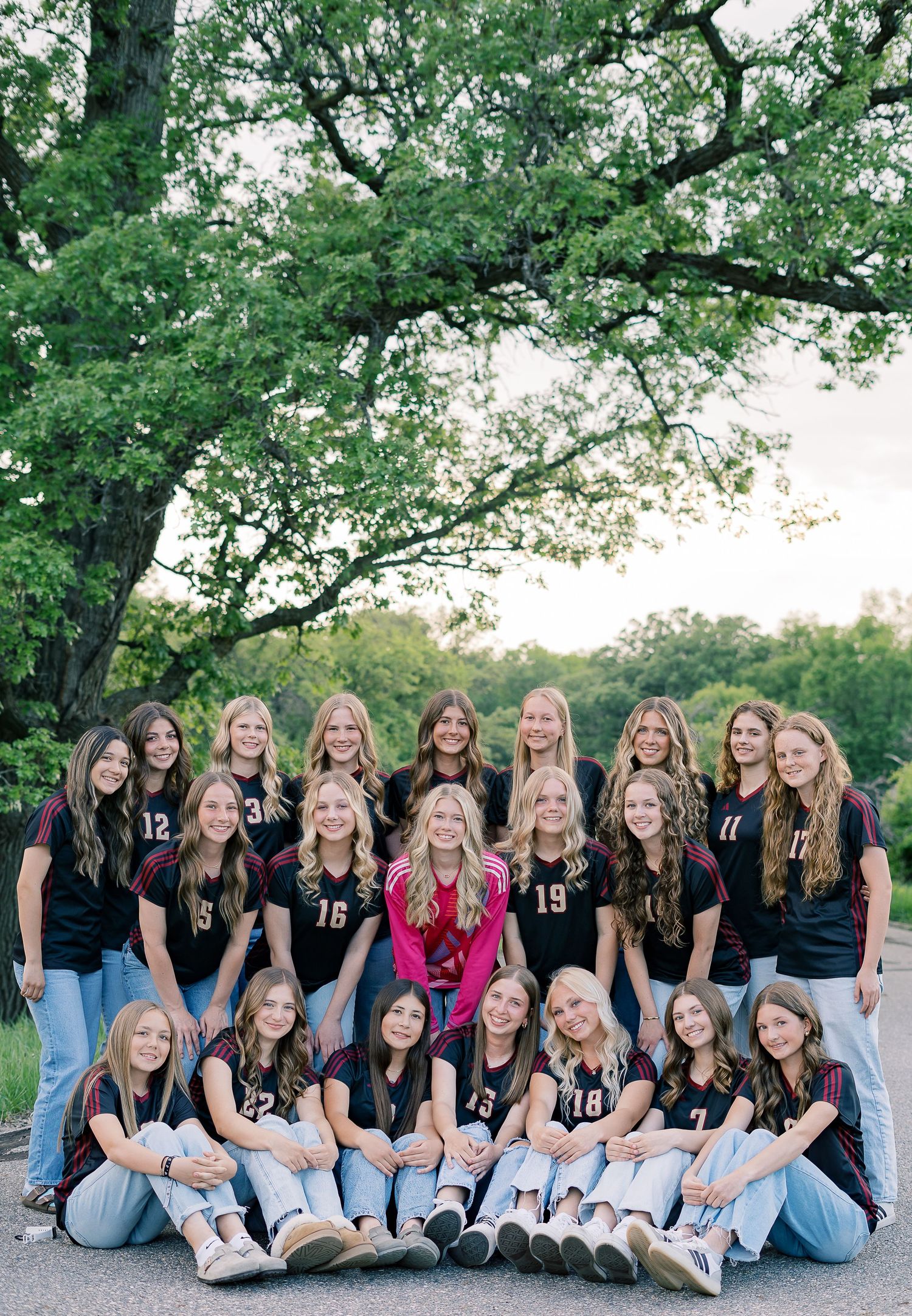 Members of a sports team pose together in matching black jerseys and blue jeans on a winding path under green trees.