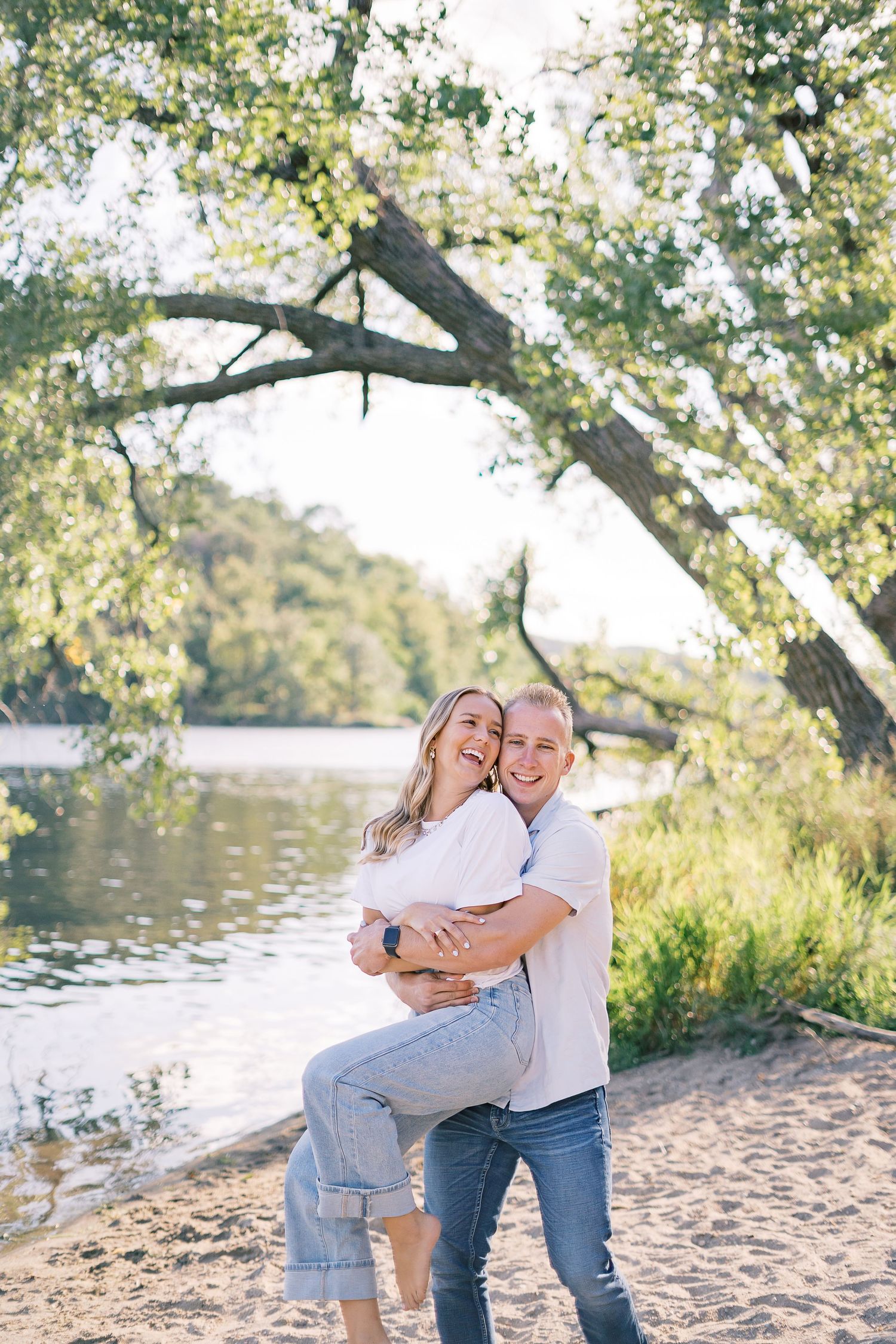 A couple shares intimate moments and laughter while sitting on a sandy beach near a lake surrounded by trees.