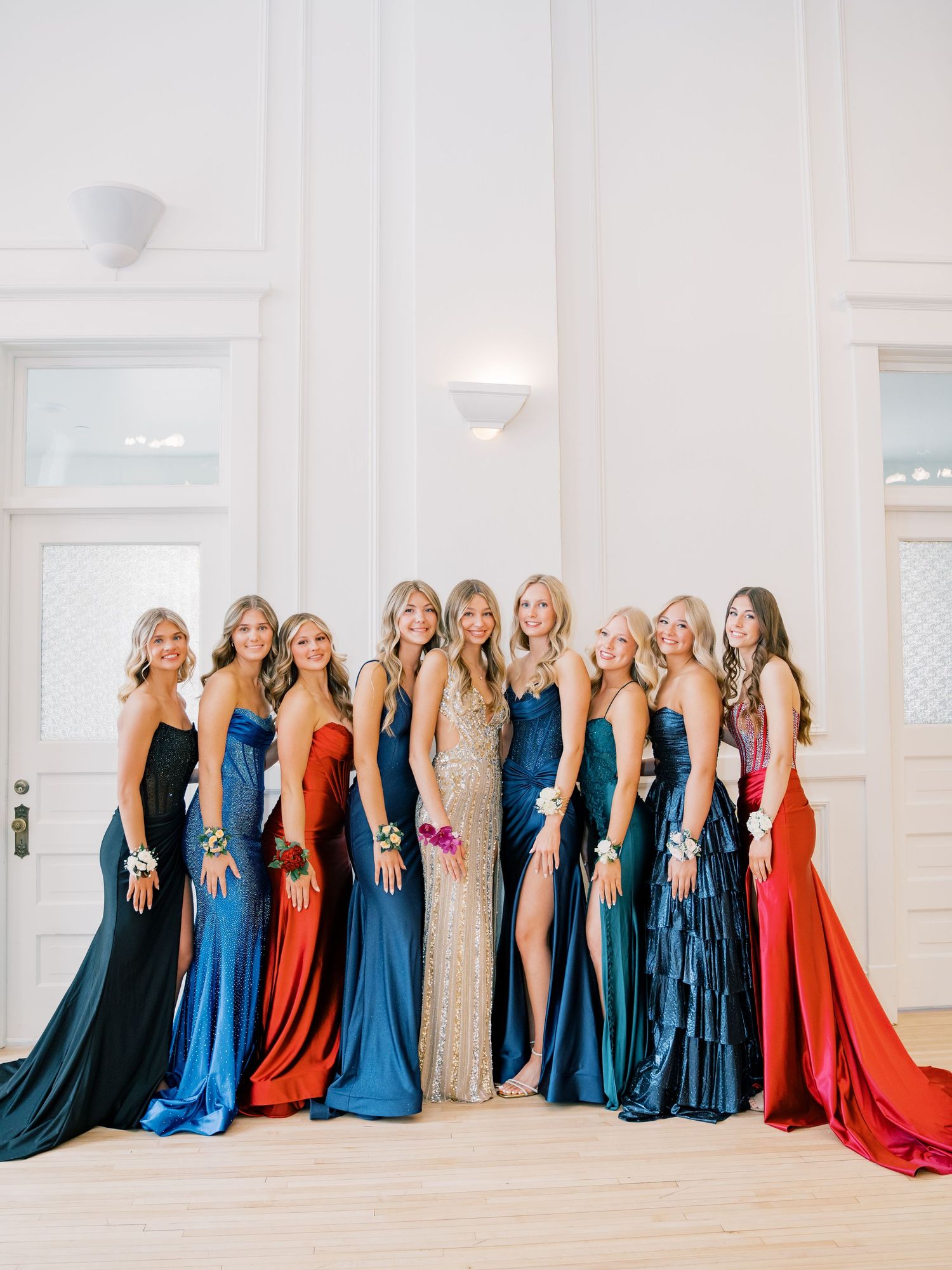 A group of nine women posing in elegant formal gowns in blue, red, and gold at a prom or formal event.