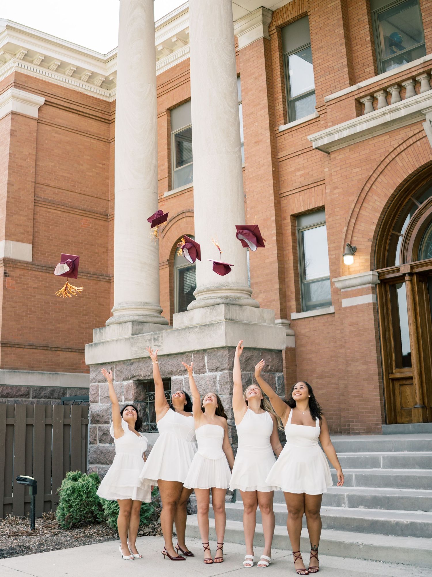 Five graduates in white dresses toss maroon caps in the air outside a brick university building.