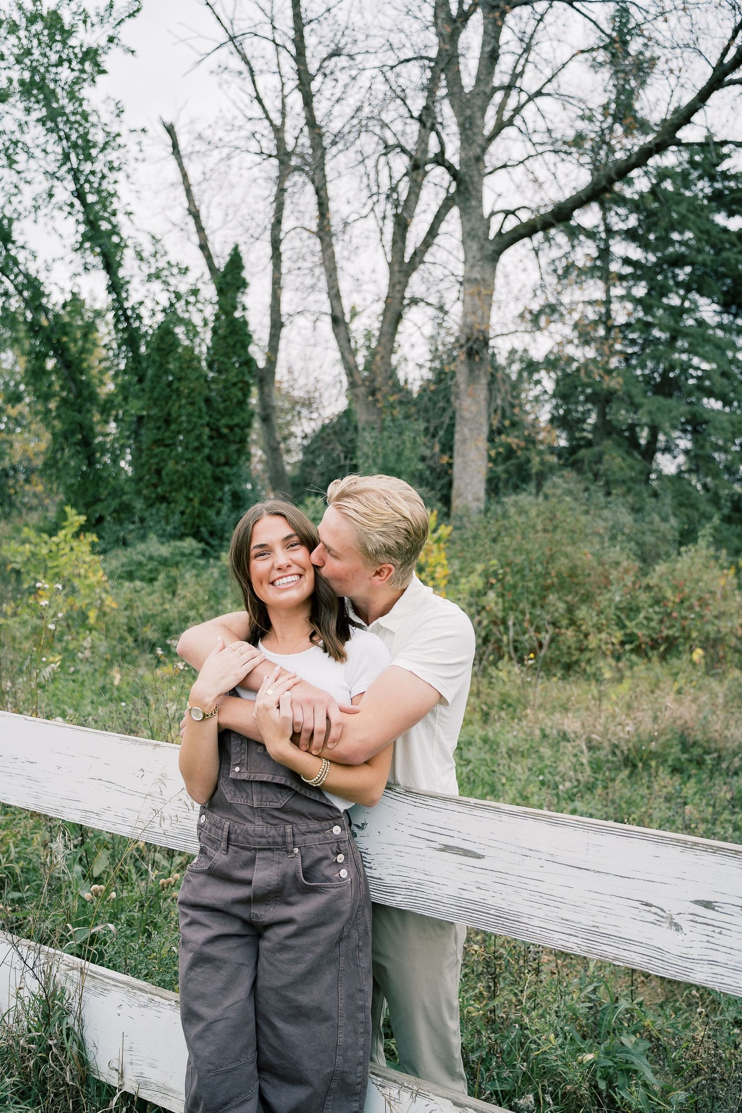 A romantic couple shares tender moments on a rustic white fence in a natural wooded setting.