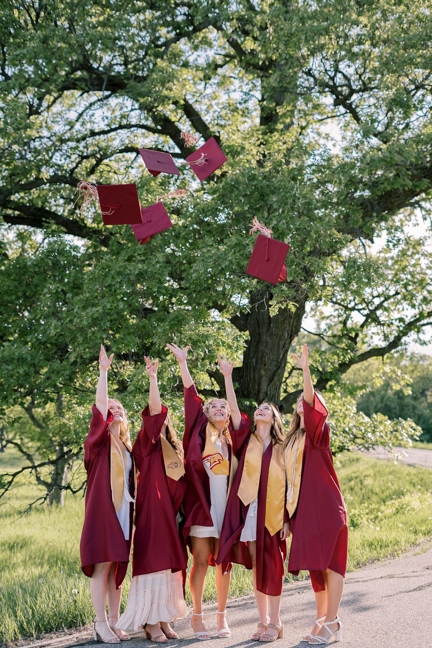 Group photos, in burgundy academic gowns celebrating their graduation outdoors on a sunny day with trees in the background.