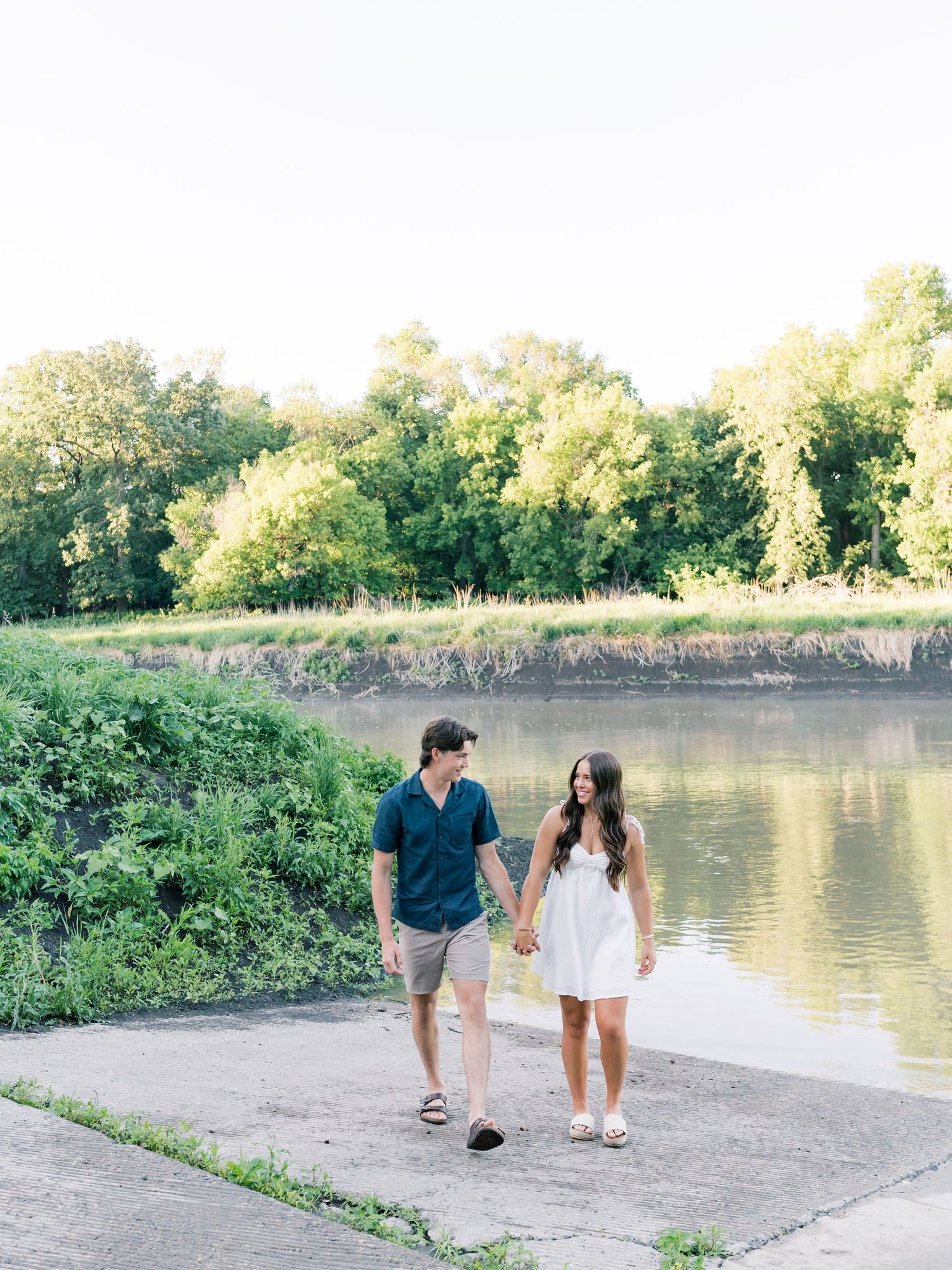 A couple holds hands and walks along a riverside path surrounded by lush green trees on a sunny day.