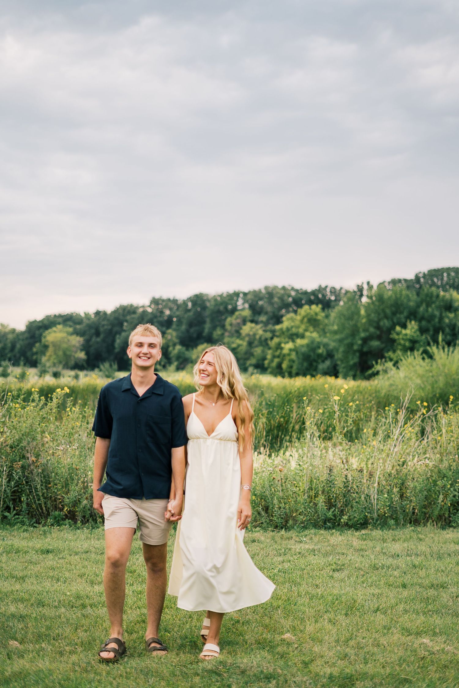 A couple walks hand in hand through a sunny summer meadow with trees in the background.