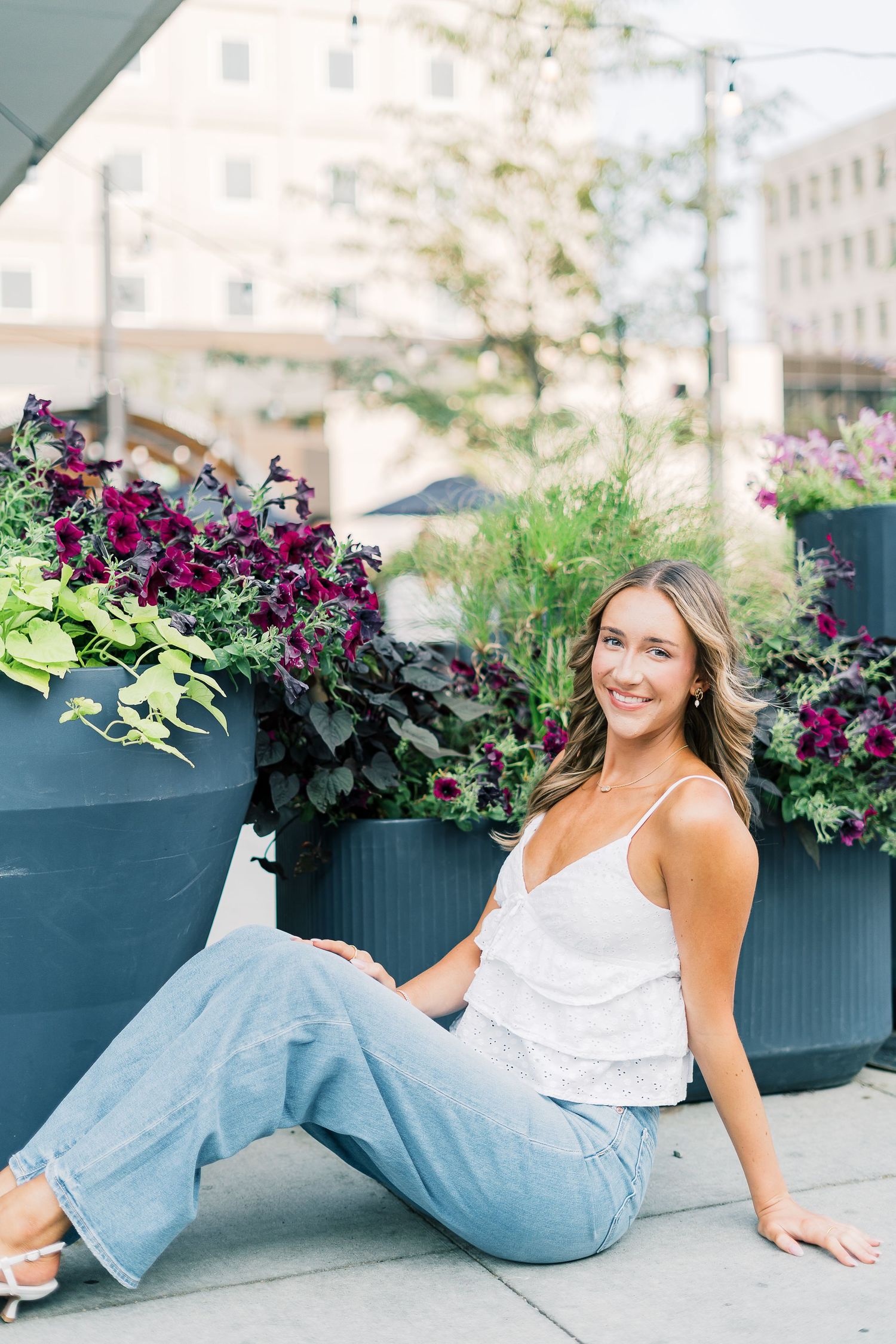 A person in jeans and white top lounges on a rooftop terrace with purple flowers in planters.