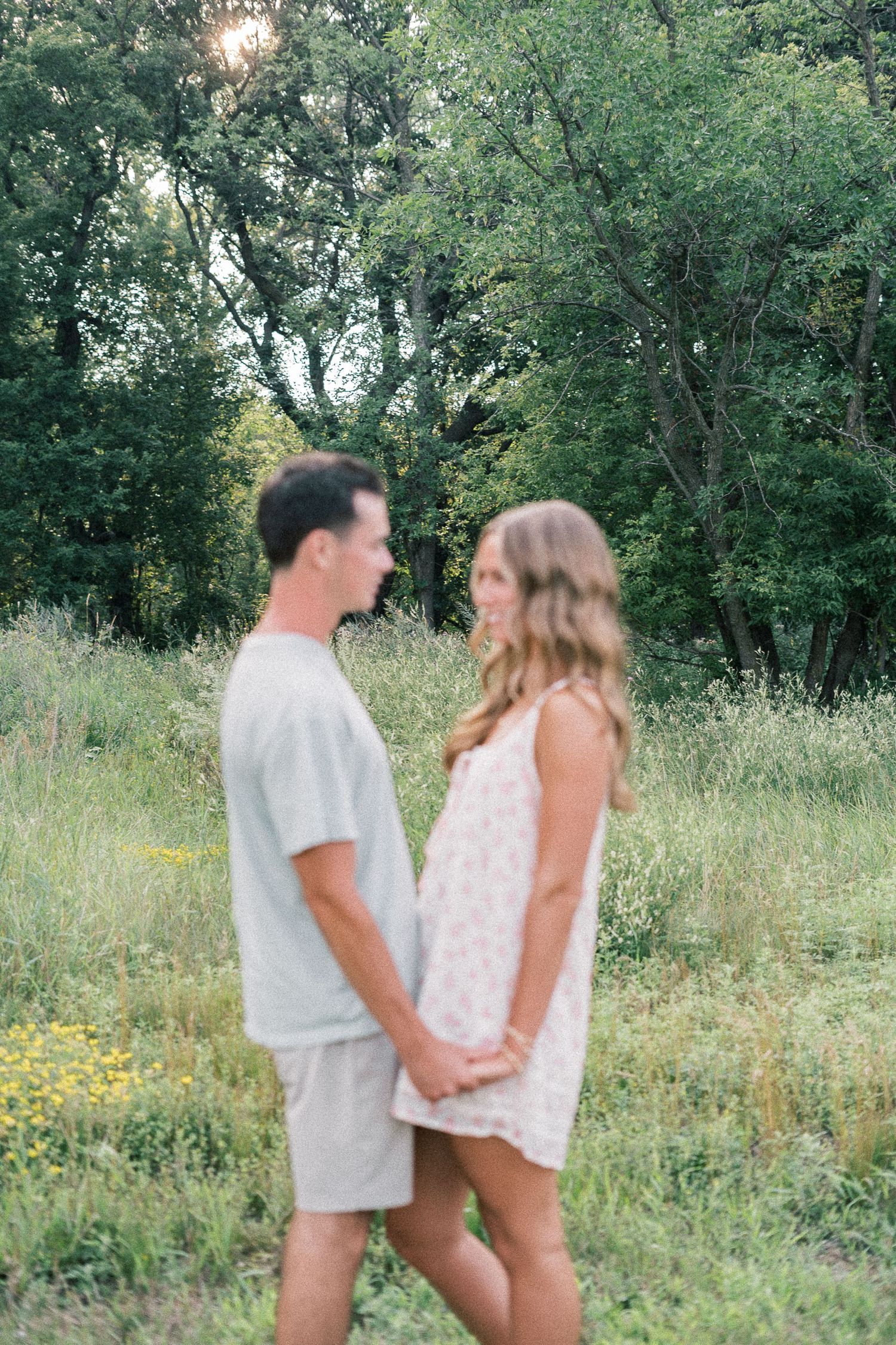 A couple embraces in a sunny meadow filled with wildflowers and trees during a romantic outdoor photoshoot.