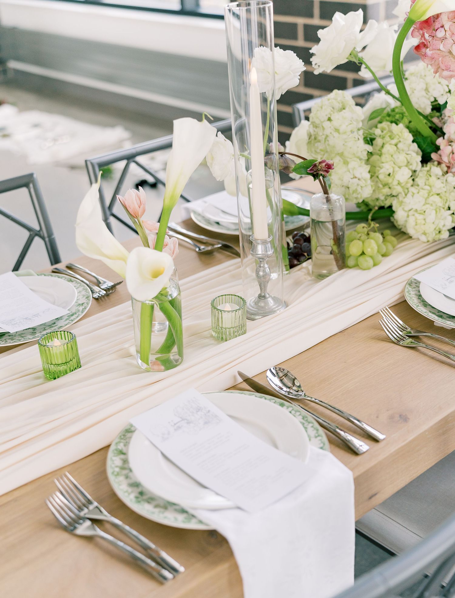 Four place settings along a rustic wooden table feature white plates, green glassware and hydrangea centerpieces.