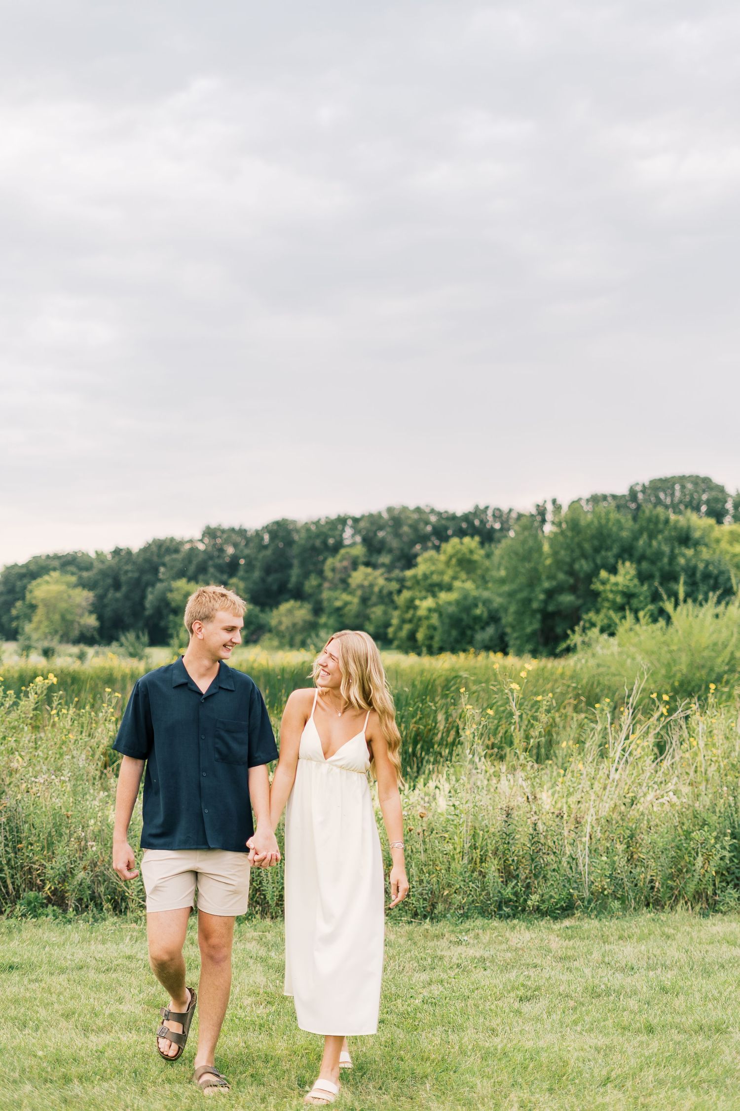 A couple walks hand in hand through a sunny meadow with wildflowers and trees in the background.
